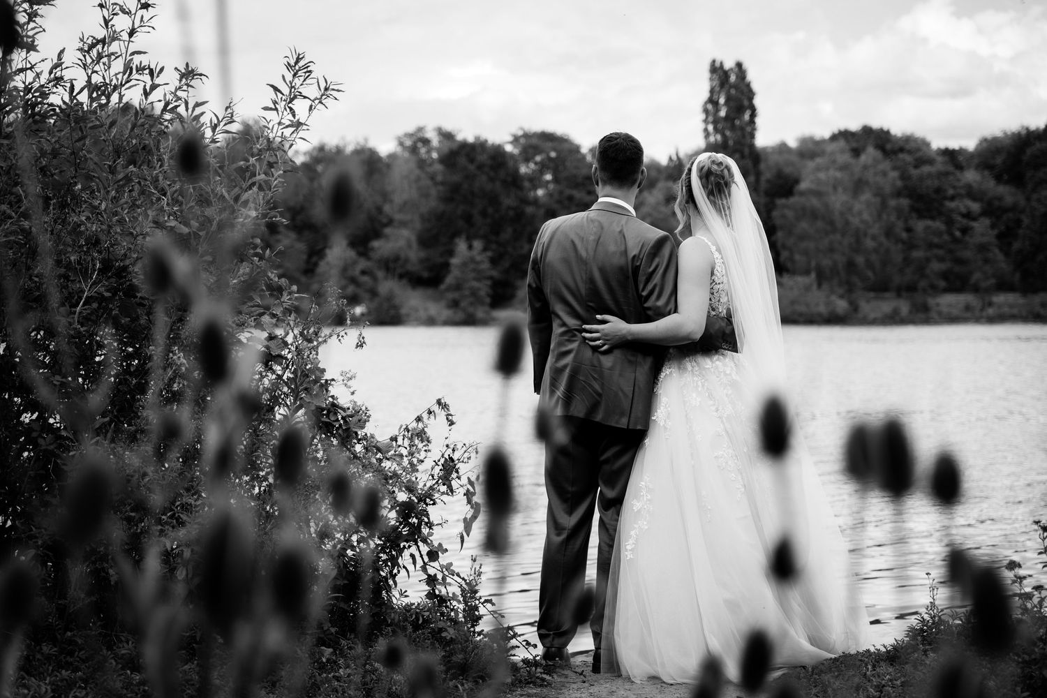 Black and white photo of a bride and groom standing by a lake, embracing and gazing at the water on their wedding day.