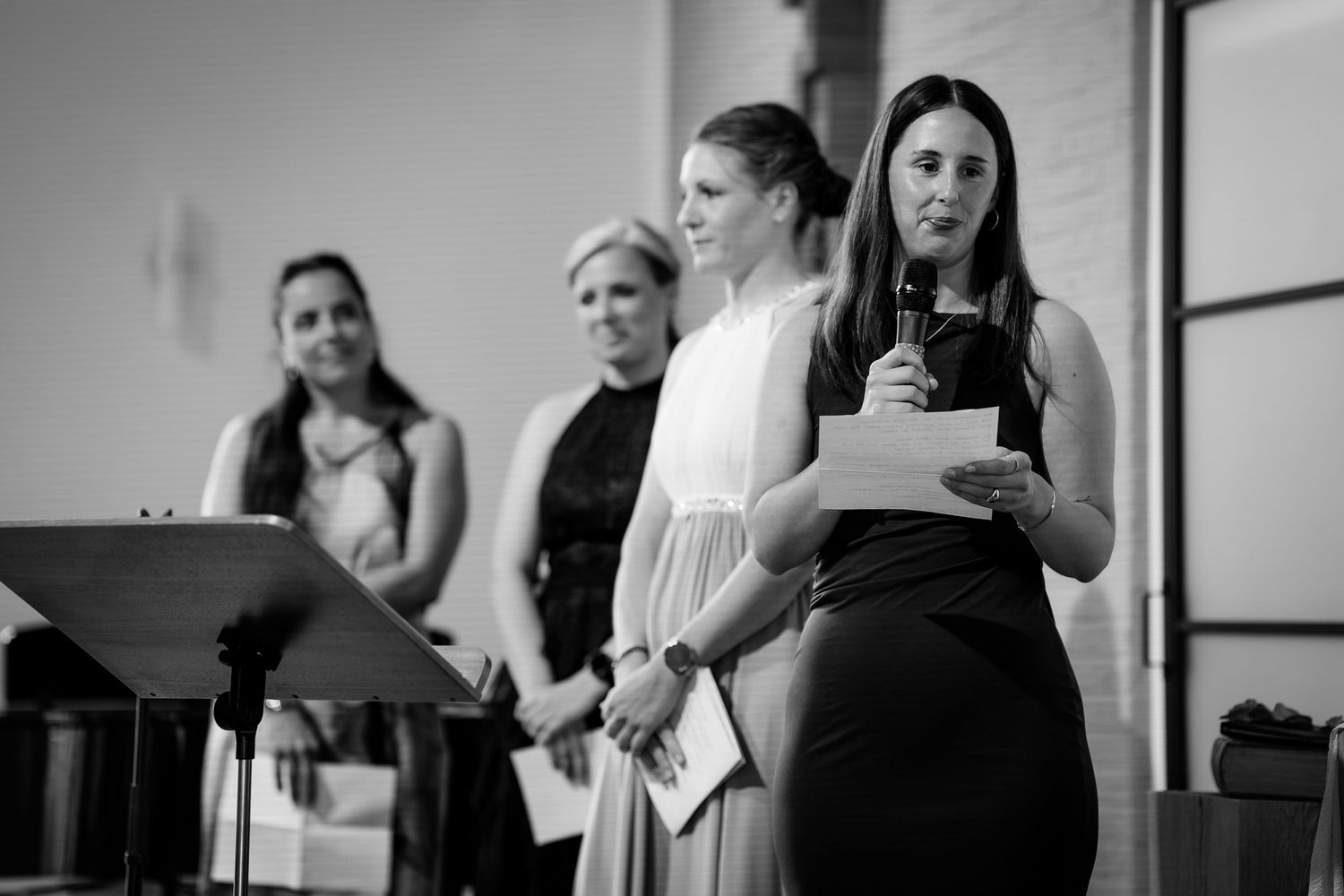 Three women stand together during a wedding reception as one reads a heartfelt speech into a microphone.