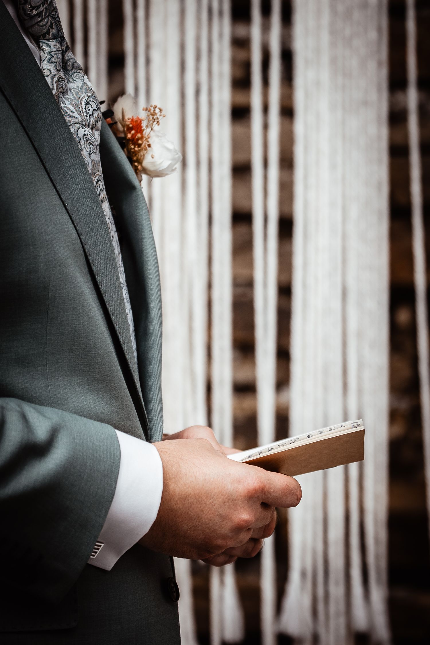 Groom in gray suit holding vow card with boutonniere at wedding ceremony.