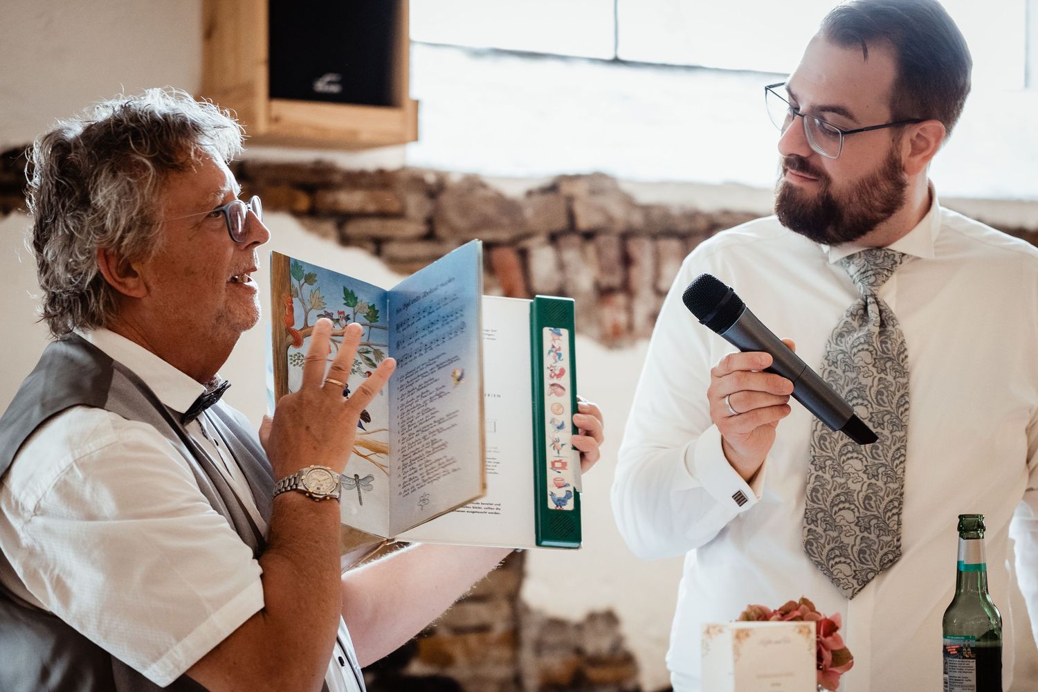 Wedding officiant reading from booklet while groom holds microphone during indoor reception speech.