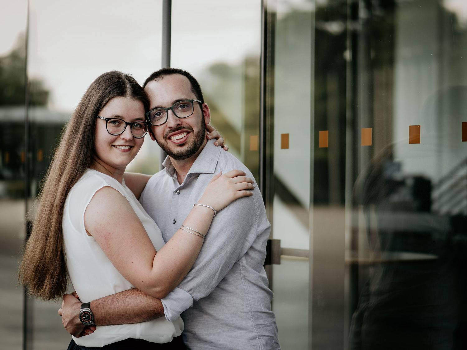 Smiling couple in glasses embracing warmly in front of modern glass building exterior.