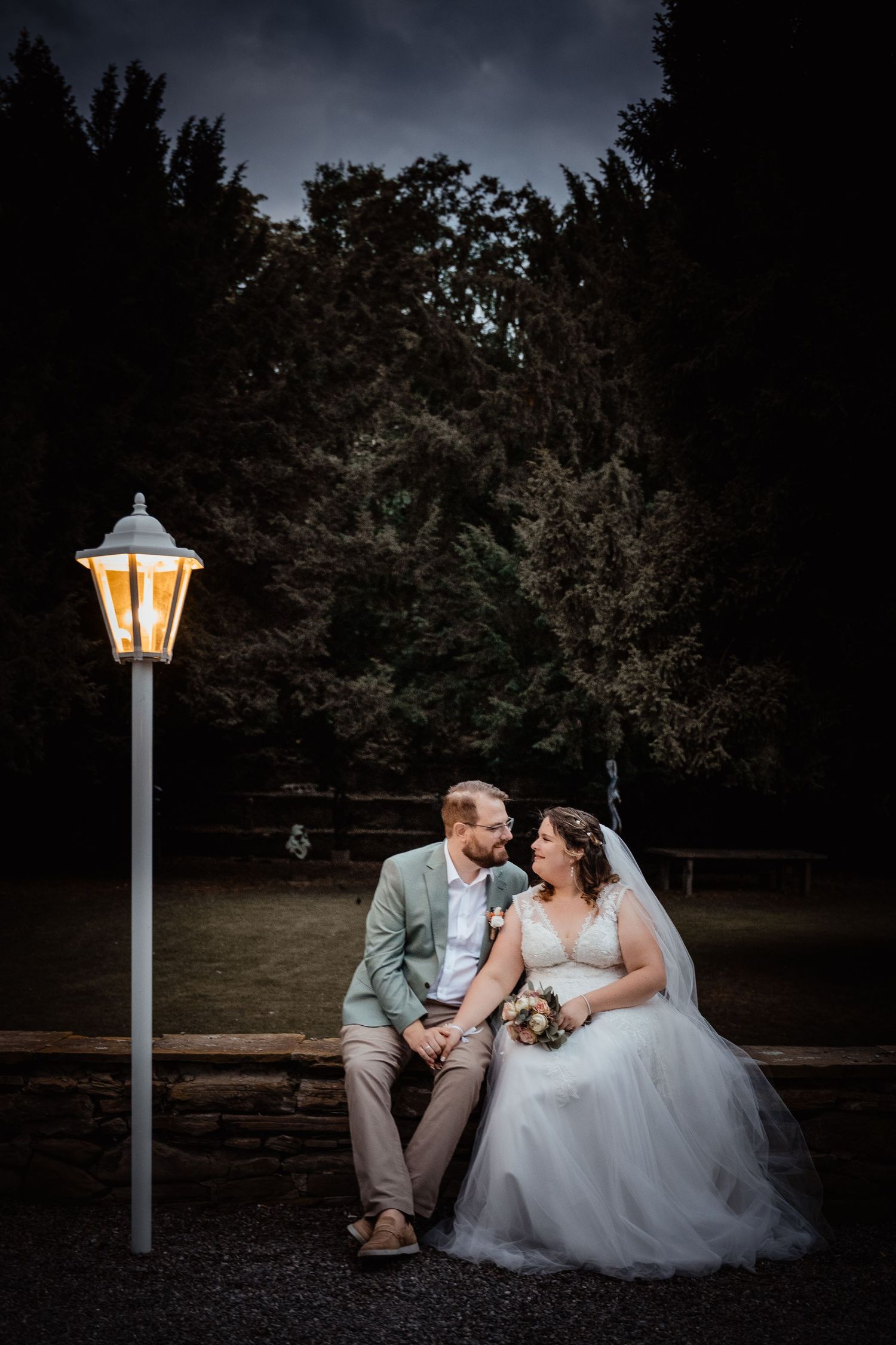 Bride and groom sit together at night near a glowing lamp post surrounded by dark trees.
