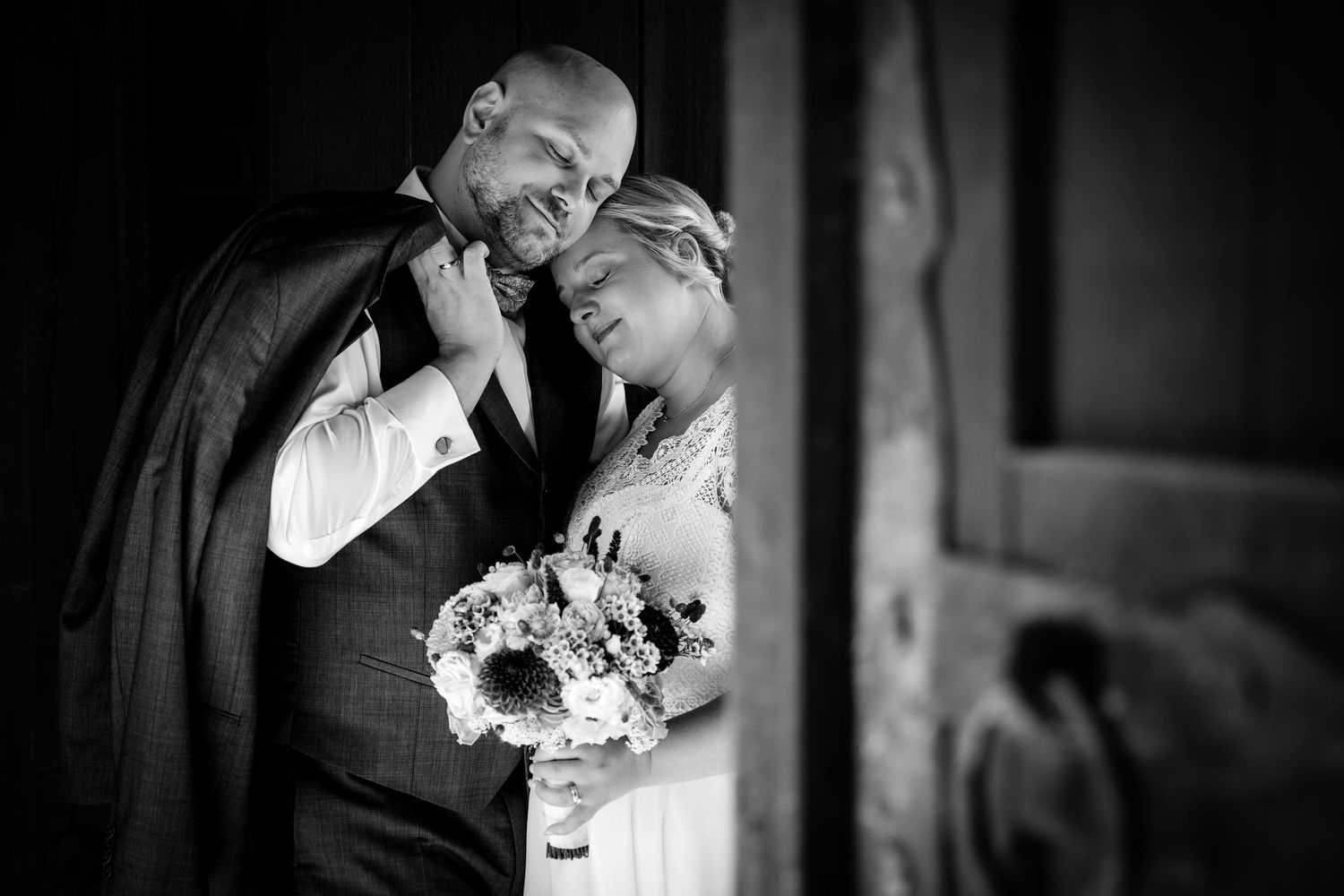 Black and white wedding photo of a smiling bride holding bouquet and groom sharing a tender moment by a wooden door.