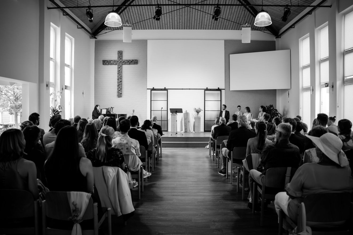 Black and white wide shot of wedding ceremony inside modern church with cross and seated guests.