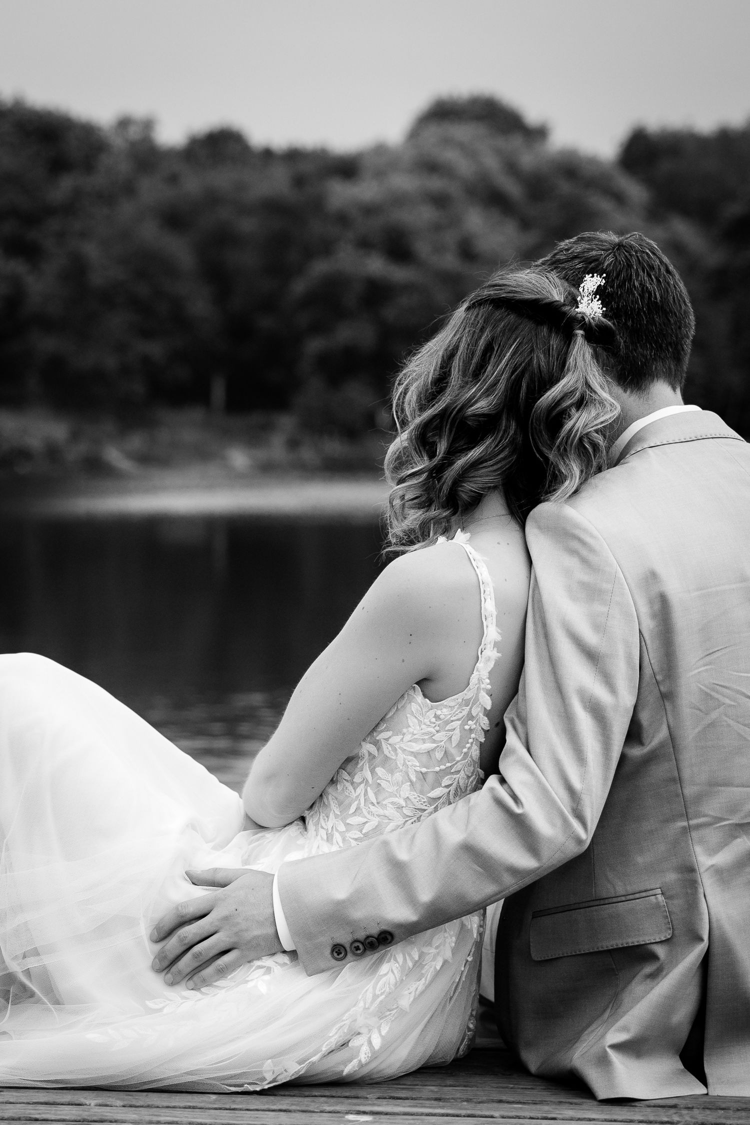 Bride and groom sitting together on a dock by a lake in a black and white photo.