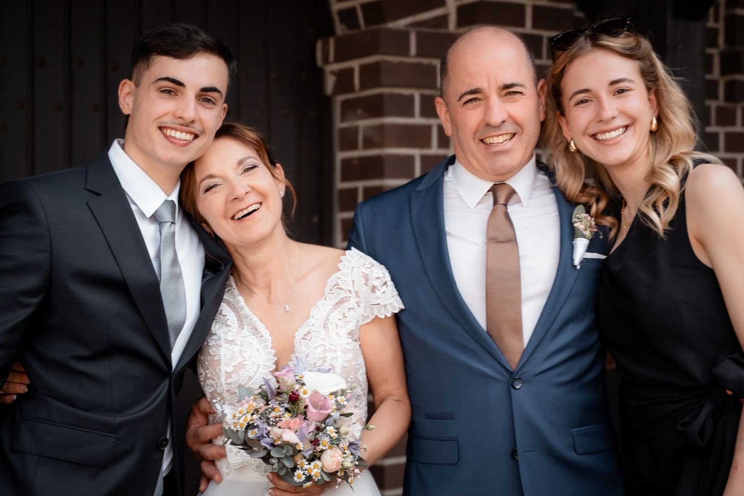 Happy bride and groom pose with two young adults outside brick building, bride holds floral bouquet.