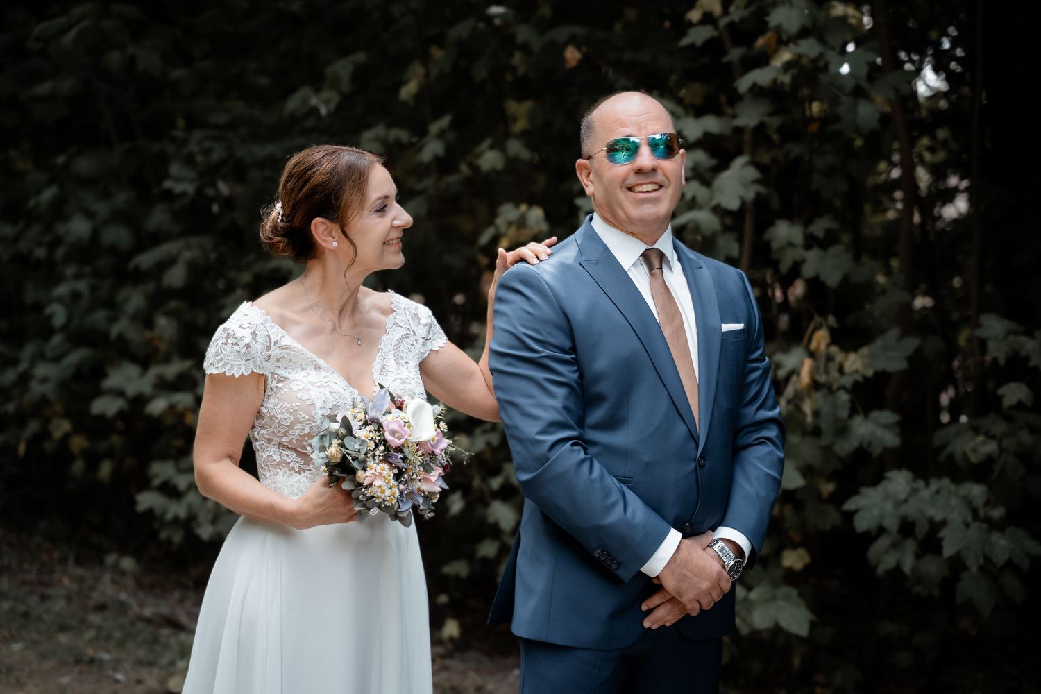 Bride holding floral bouquet stands with groom in blue suit and sunglasses outdoors before wedding ceremony.