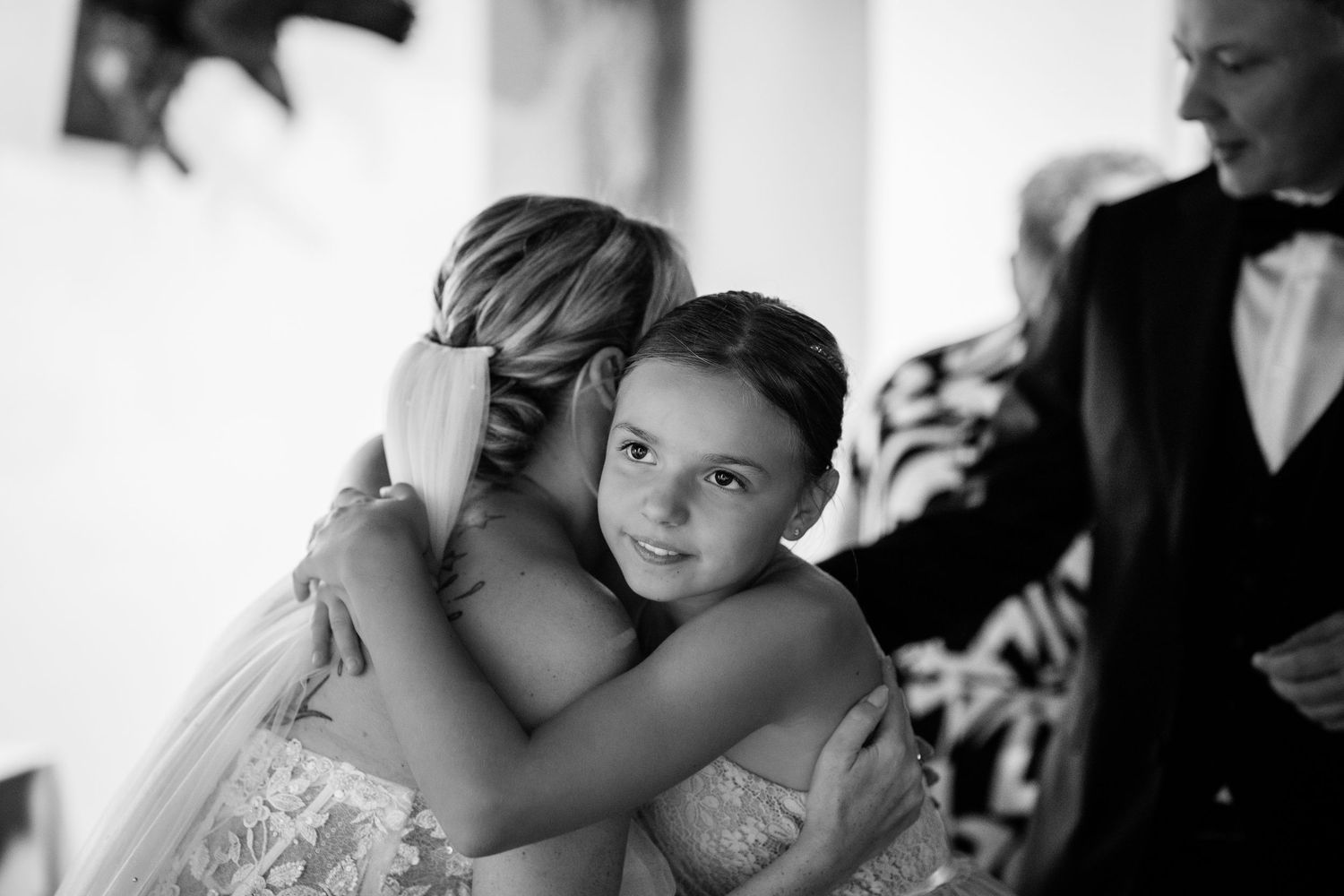 Black and white photo of a young girl hugging a bride in a lace wedding dress at a wedding ceremony.