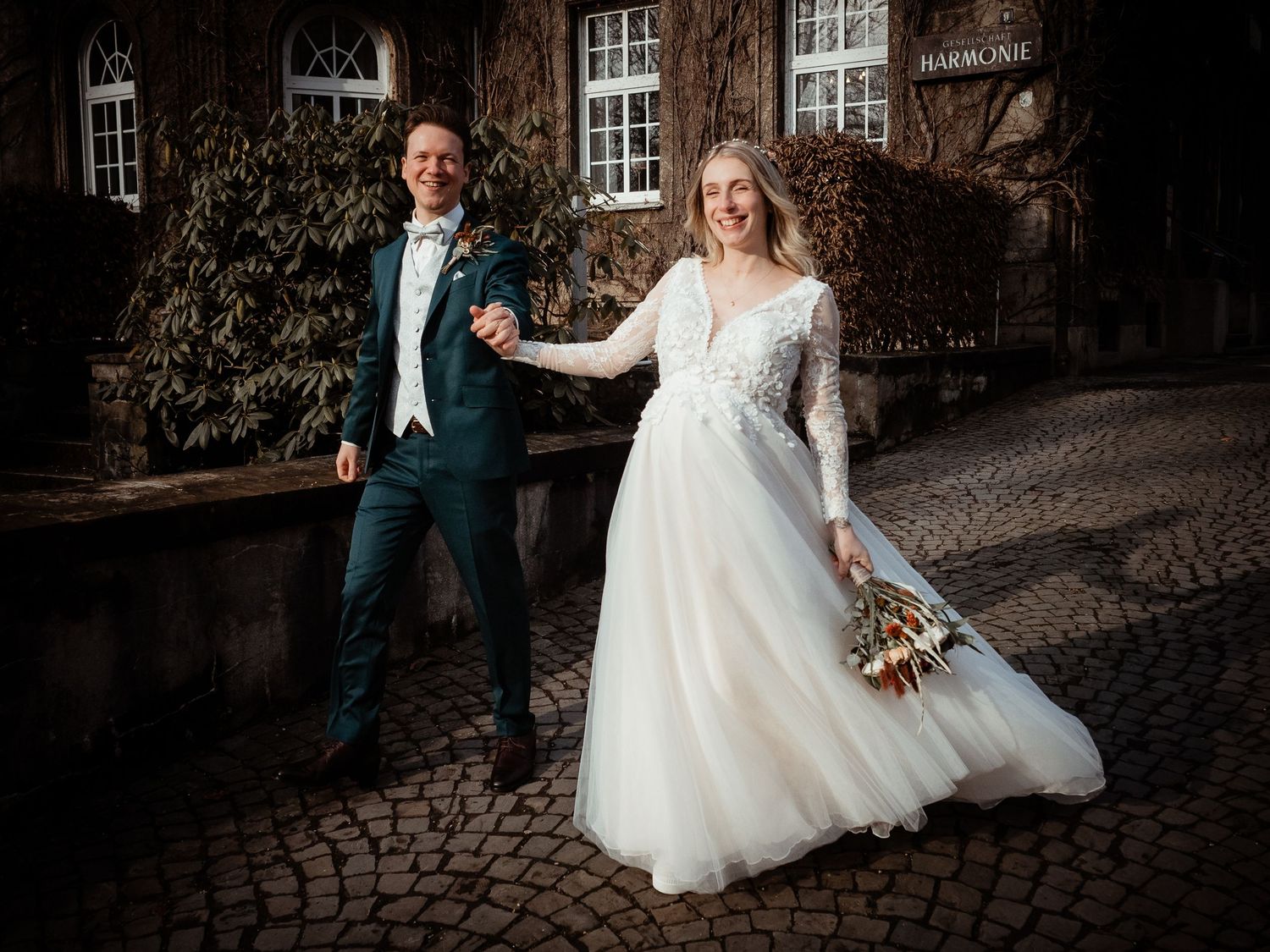 Happy bride and groom holding hands and laughing outside a historic ivy-covered building on cobblestones.