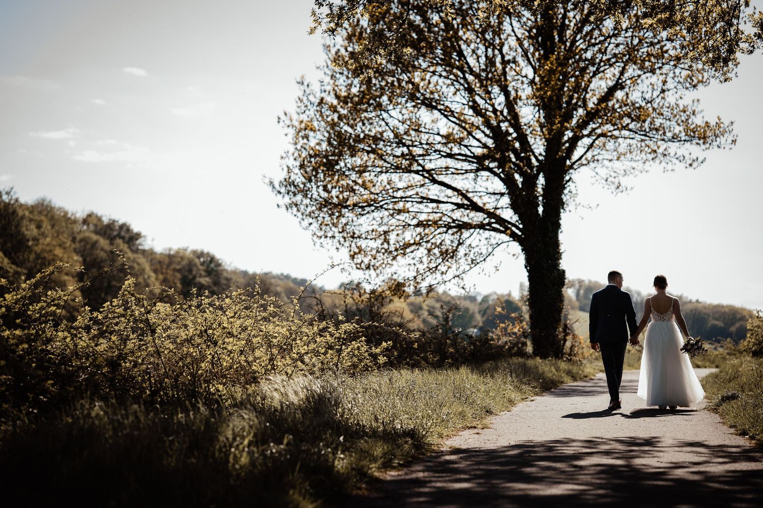 A couple walks down a tree-lined path in a romantic outdoor setting during golden hour.