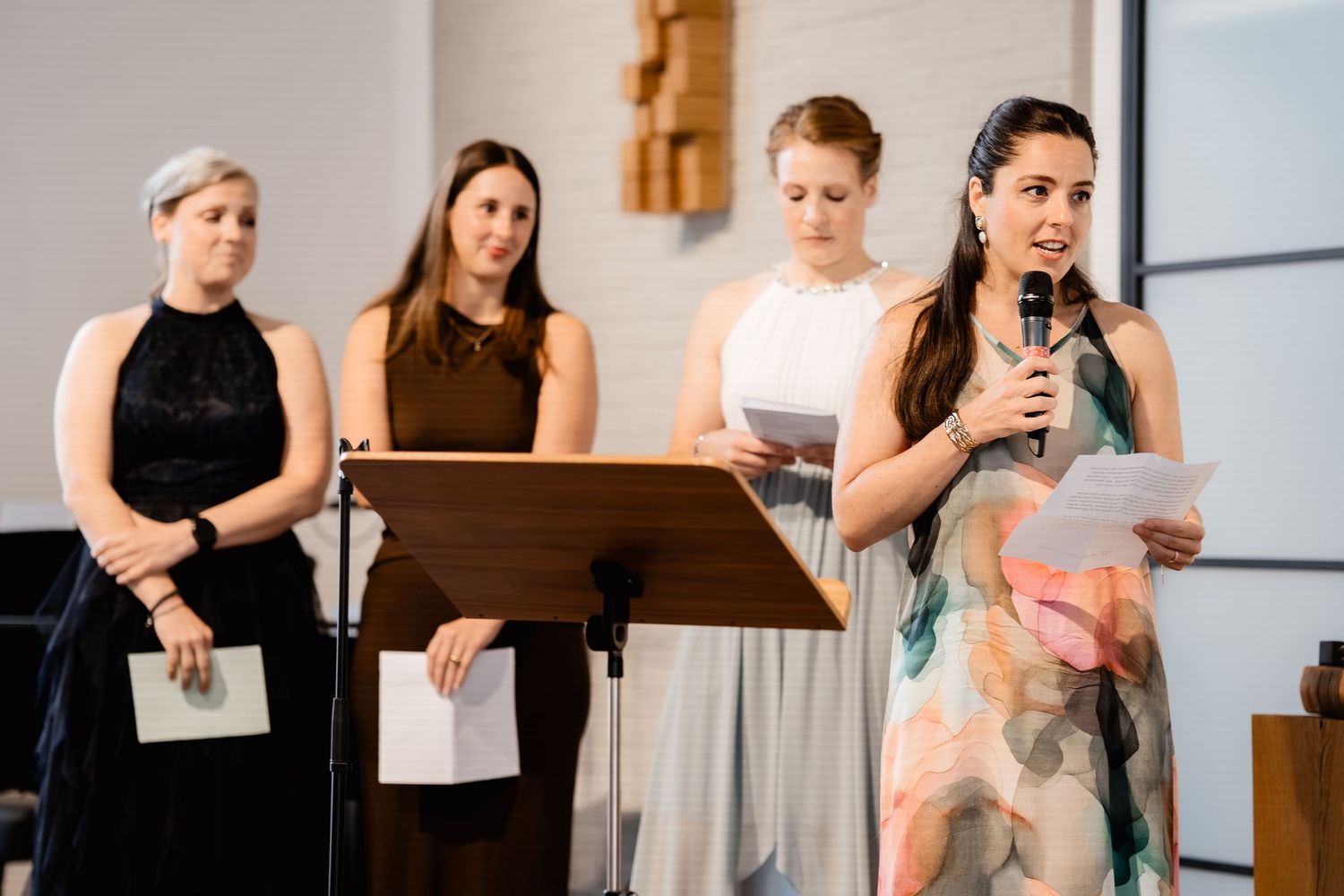 Four women stand at a podium during a wedding ceremony, one speaking into a microphone while holding papers.