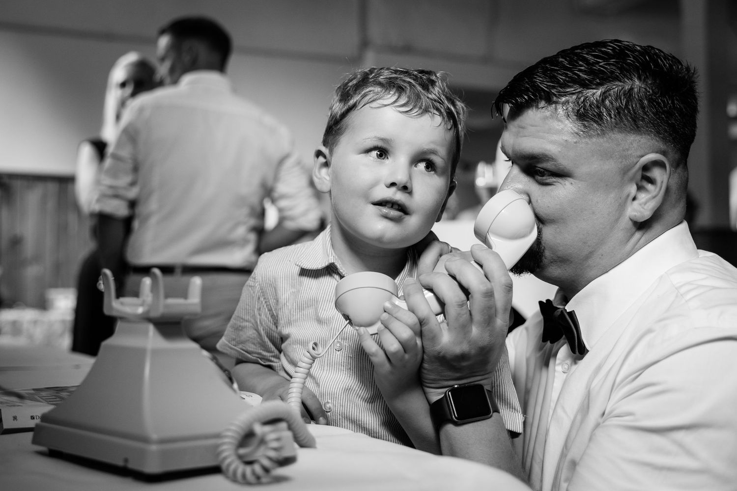 Black and white photo of a young boy and man playing with a vintage toy phone at a wedding reception.