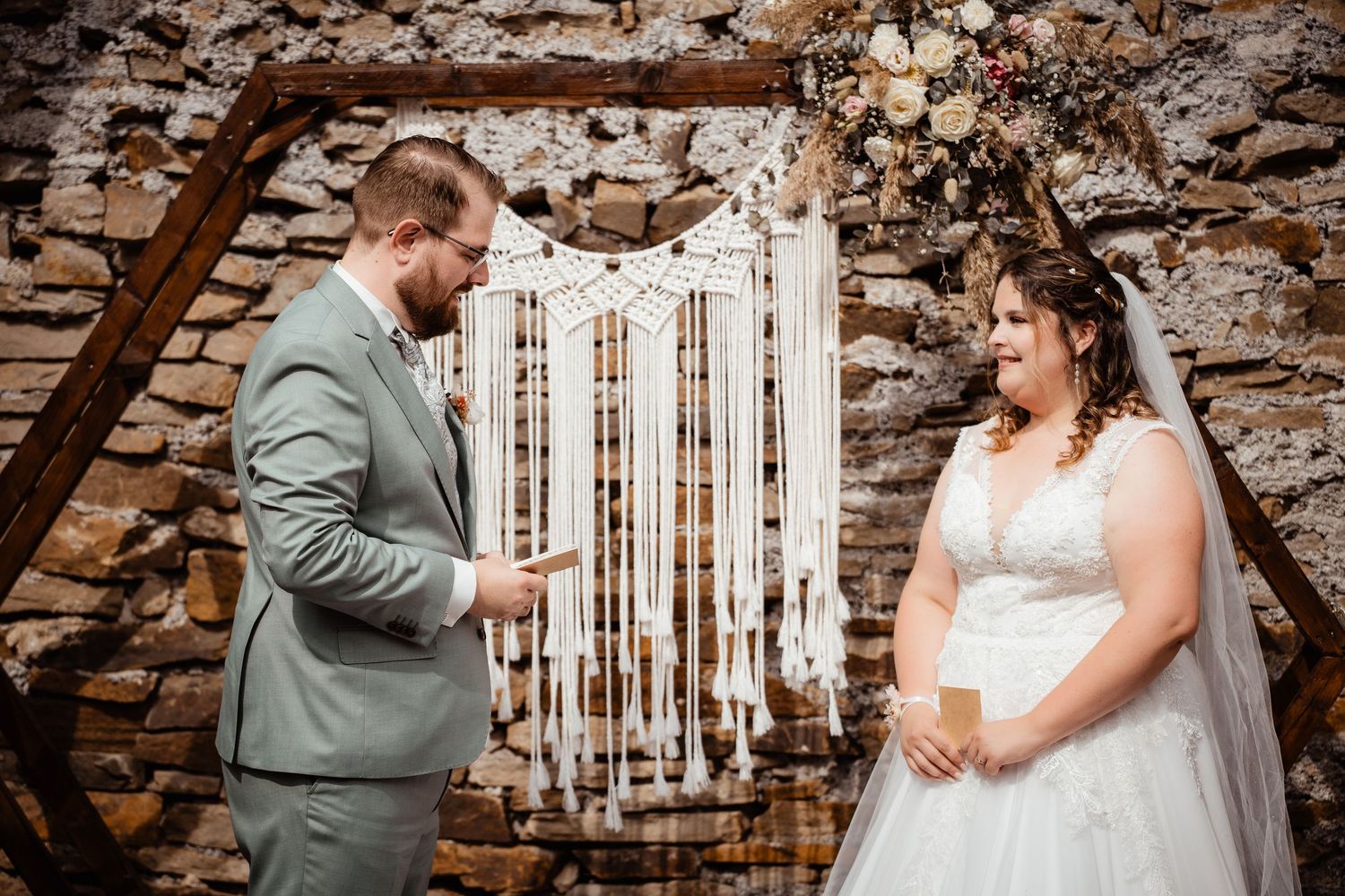 Bride and groom exchange vows before rustic hexagonal arch with macrame backdrop and stone wall.