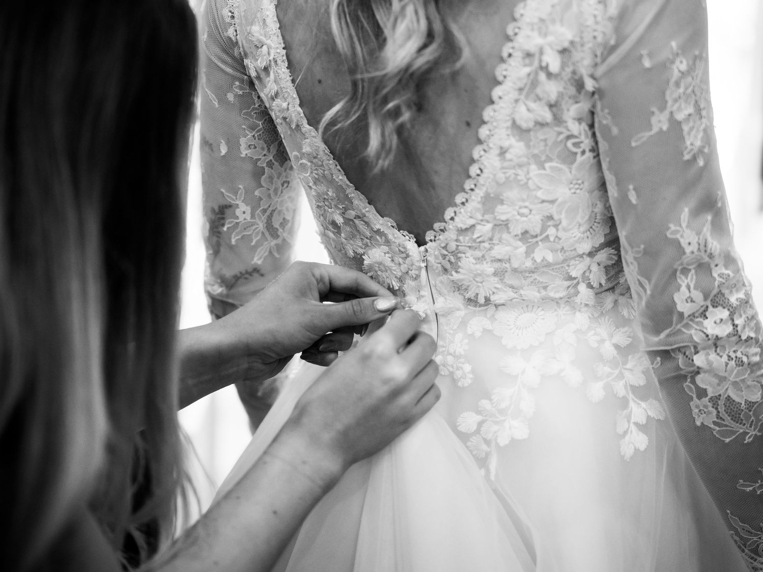 Black and white photo of a bride having her lace wedding dress buttoned up from behind by a bridesmaid.