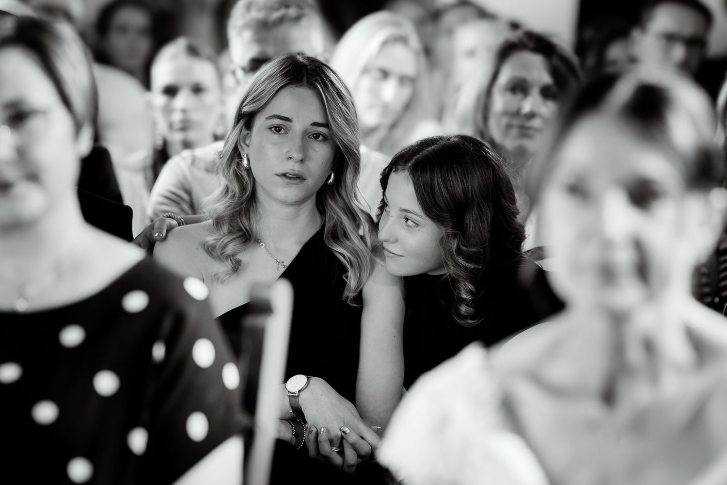 Black and white photo of wedding guests watching ceremony, two young women in foreground looking on.