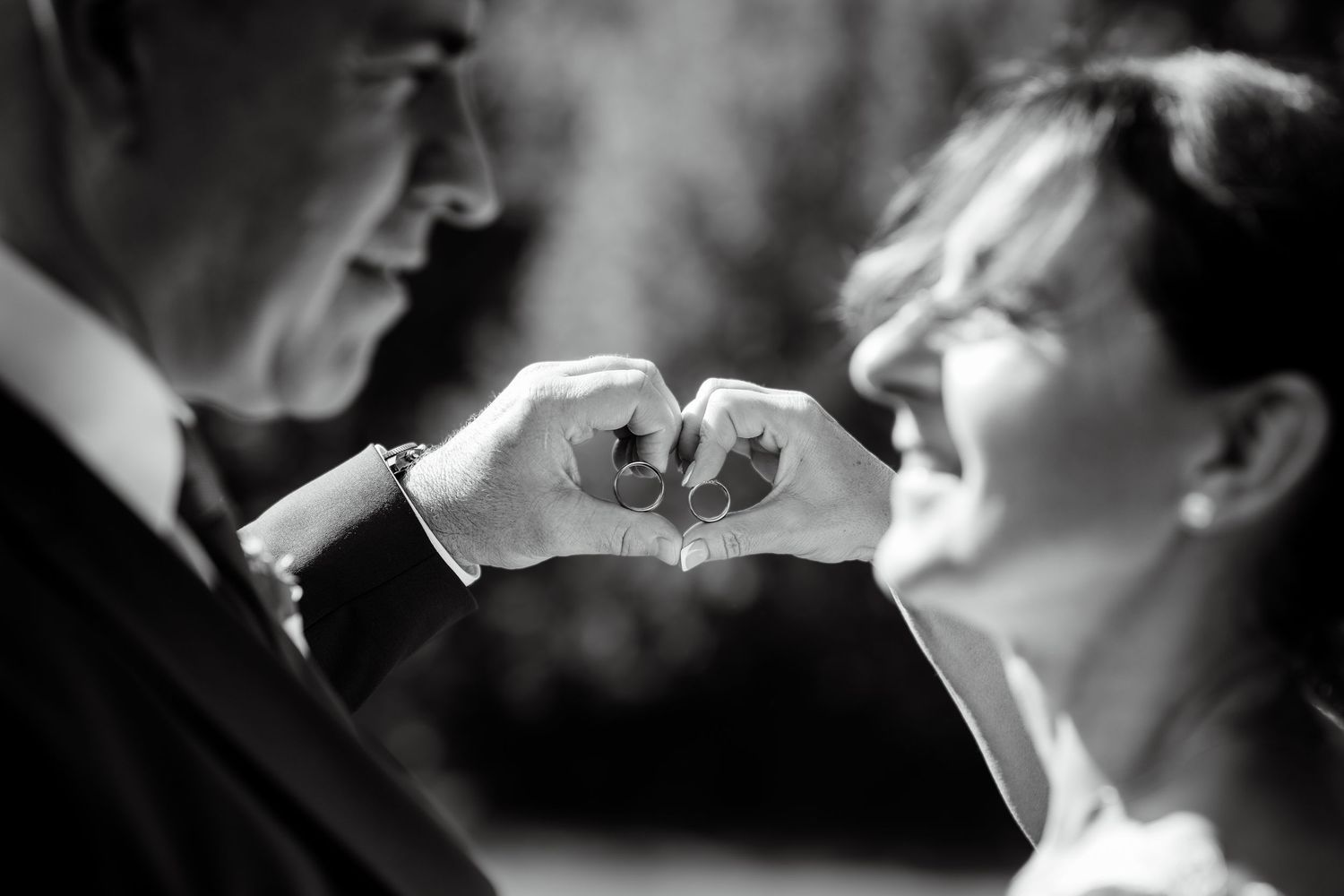 Black and white close-up of newlyweds making a heart shape with their hands, both smiling joyfully.