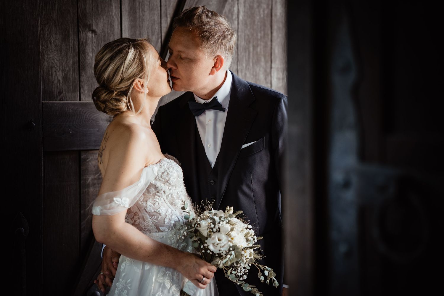 Bride and groom sharing a tender kiss against rustic dark wooden door.