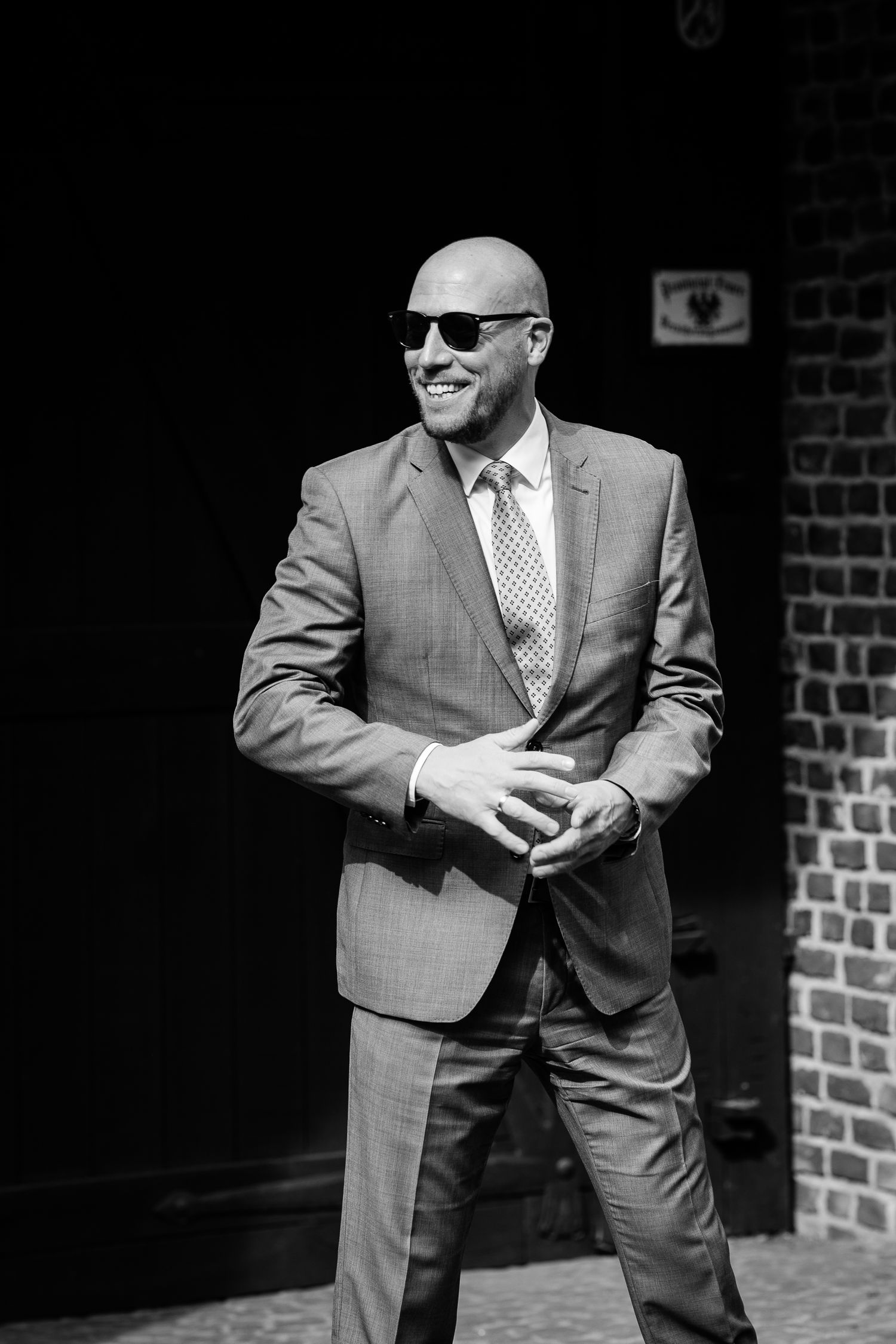 Bald man in a tailored suit and sunglasses smiling outdoors near a brick wall, black and white photo.