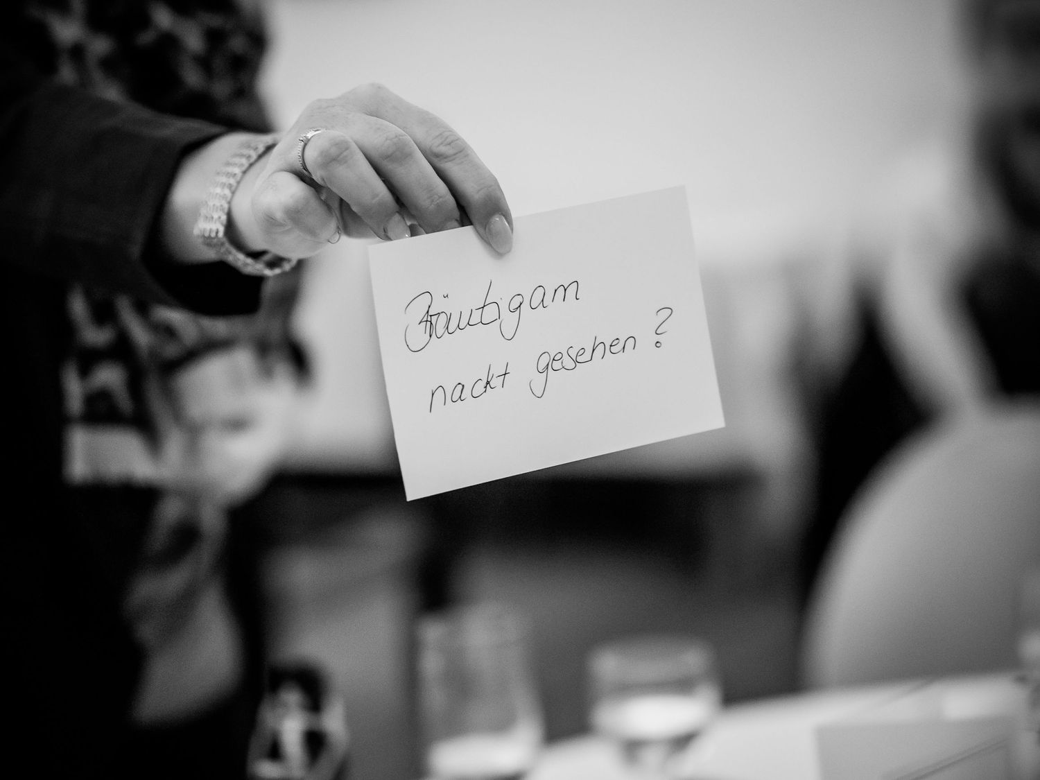 Hand holding a card with German text 'Bräutigam nackt gesehen?' at a wedding event in black and white.