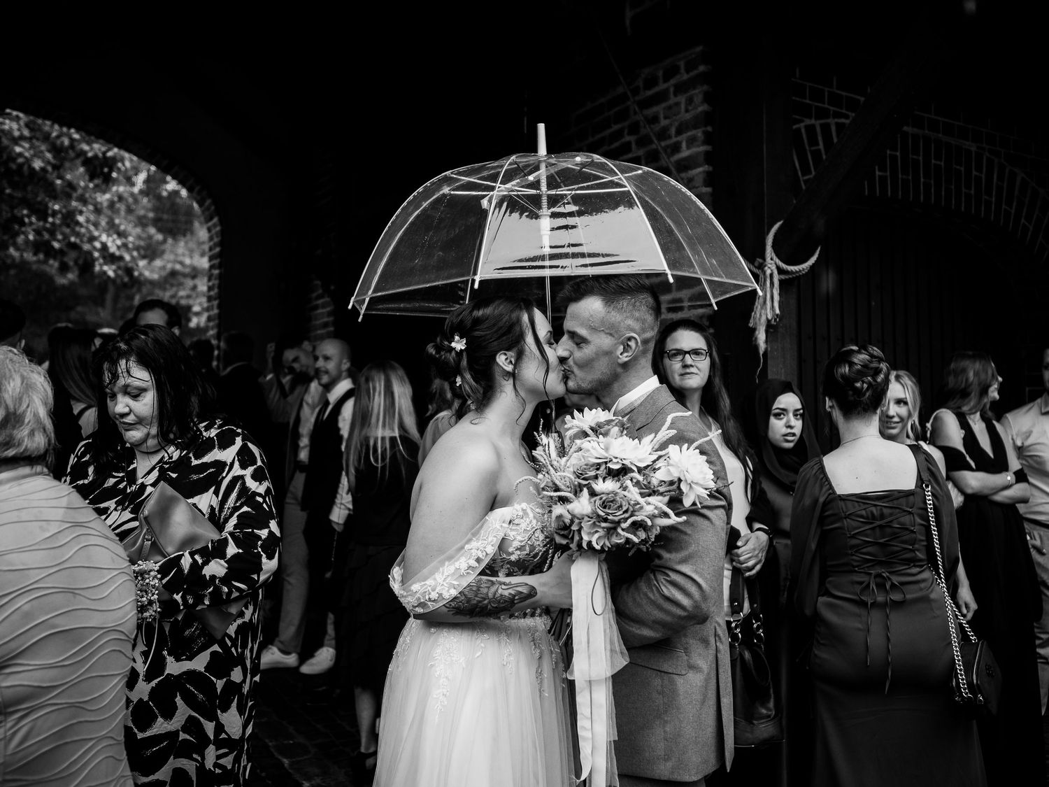 Bride and groom share a kiss under a clear umbrella while guests celebrate around them at an outdoor wedding.