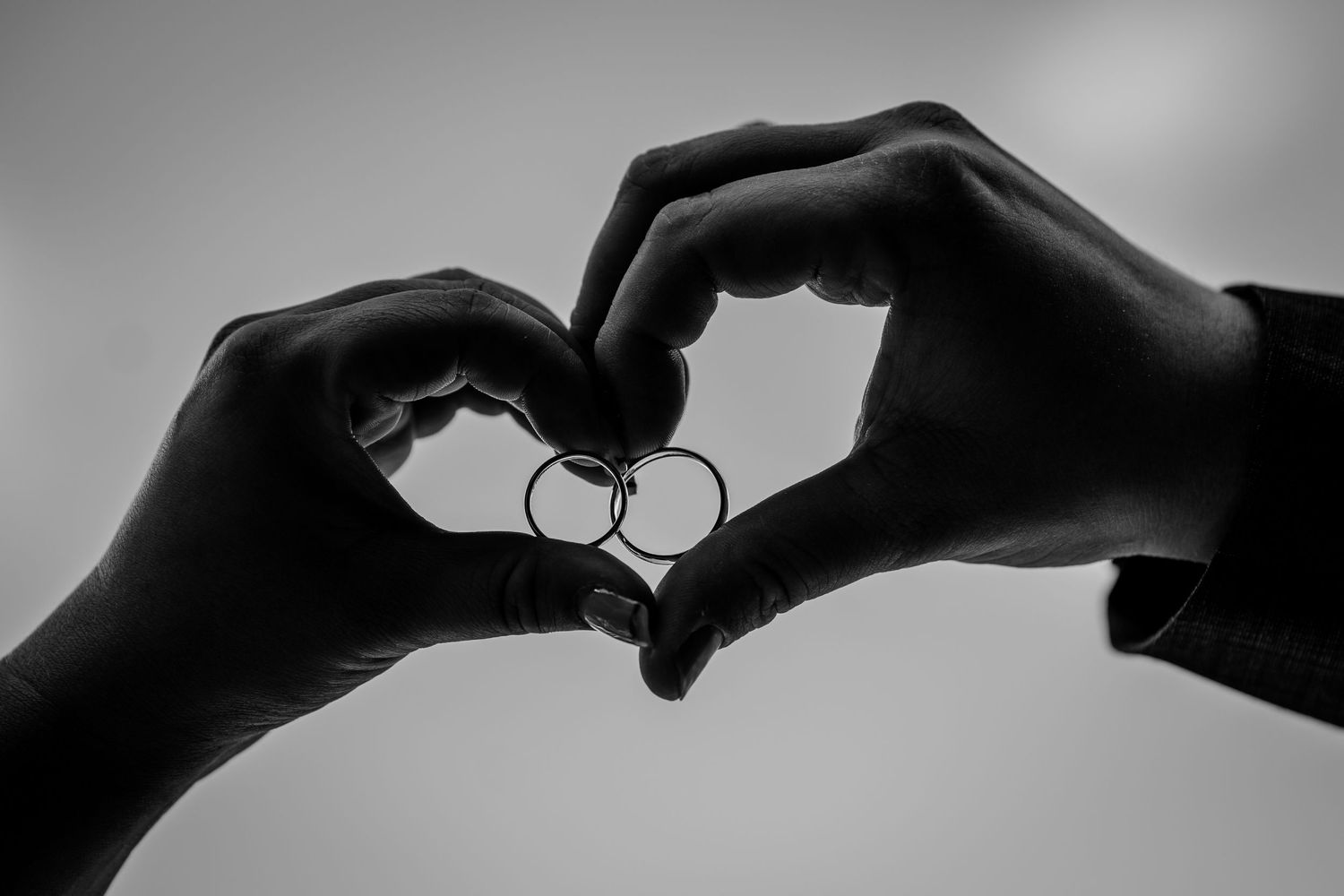 Black and white photo of two hands forming a heart shape around two interlocked wedding rings against a soft sky.