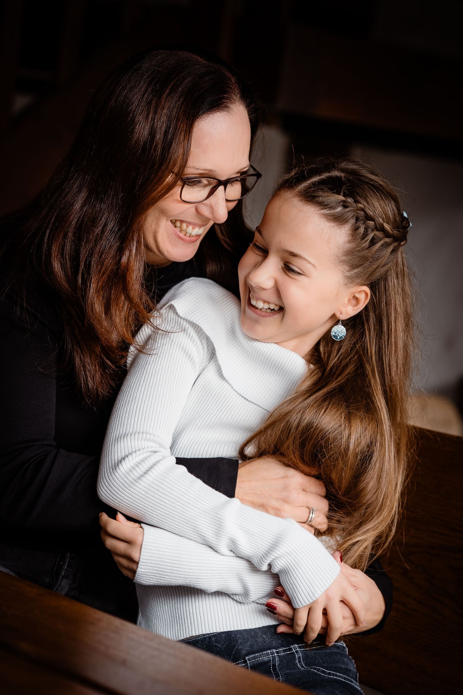 A mother and daughter share a joyful hug and laugh together, the girl in a white top with braided hair.