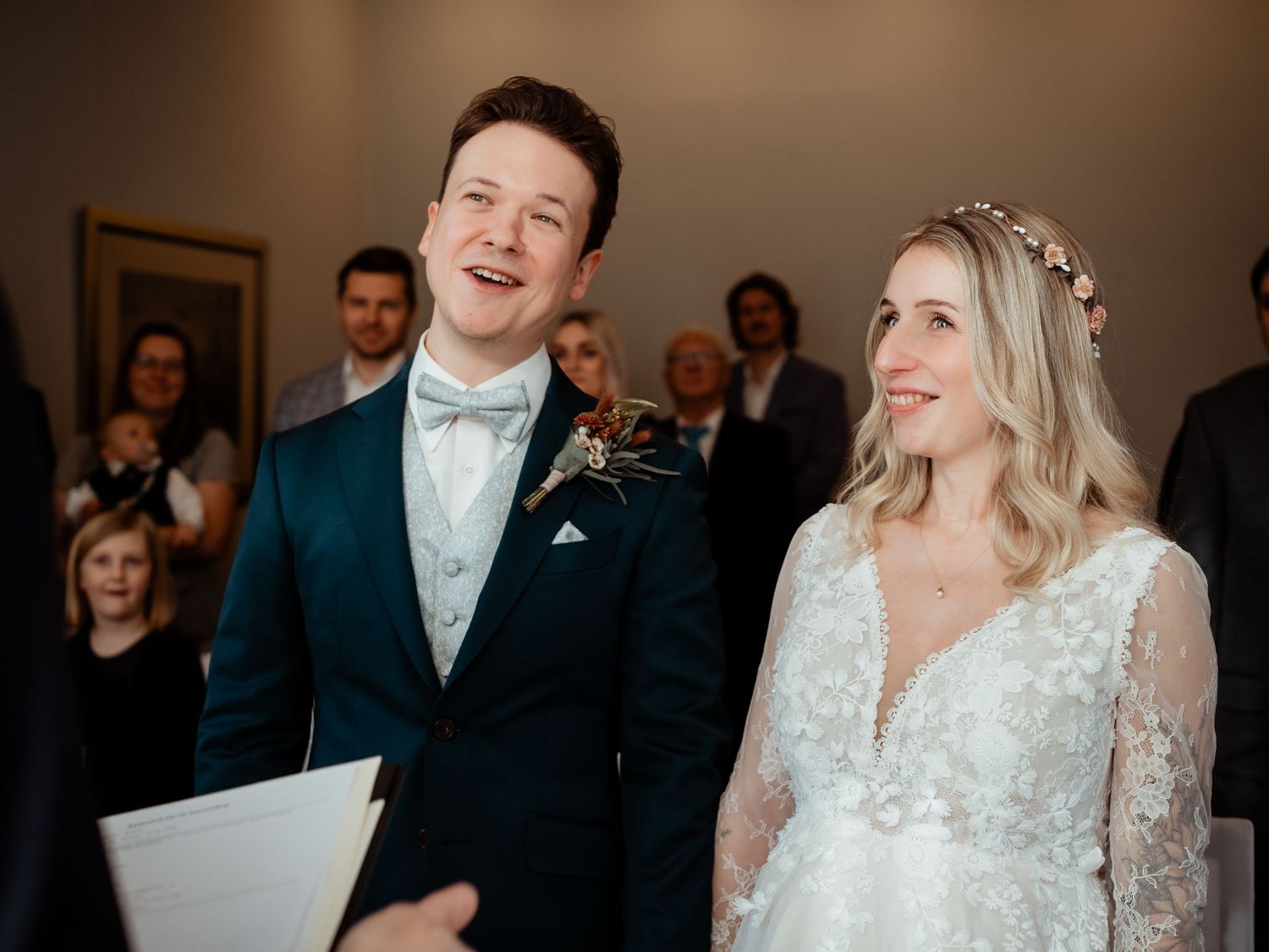 Joyful bride and groom laughing during their wedding ceremony surrounded by guests in a bright venue.