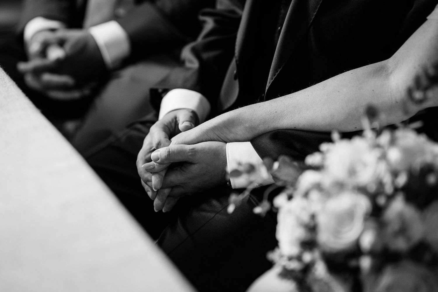 Black and white close-up of bride and groom holding hands during wedding ceremony with bouquet.