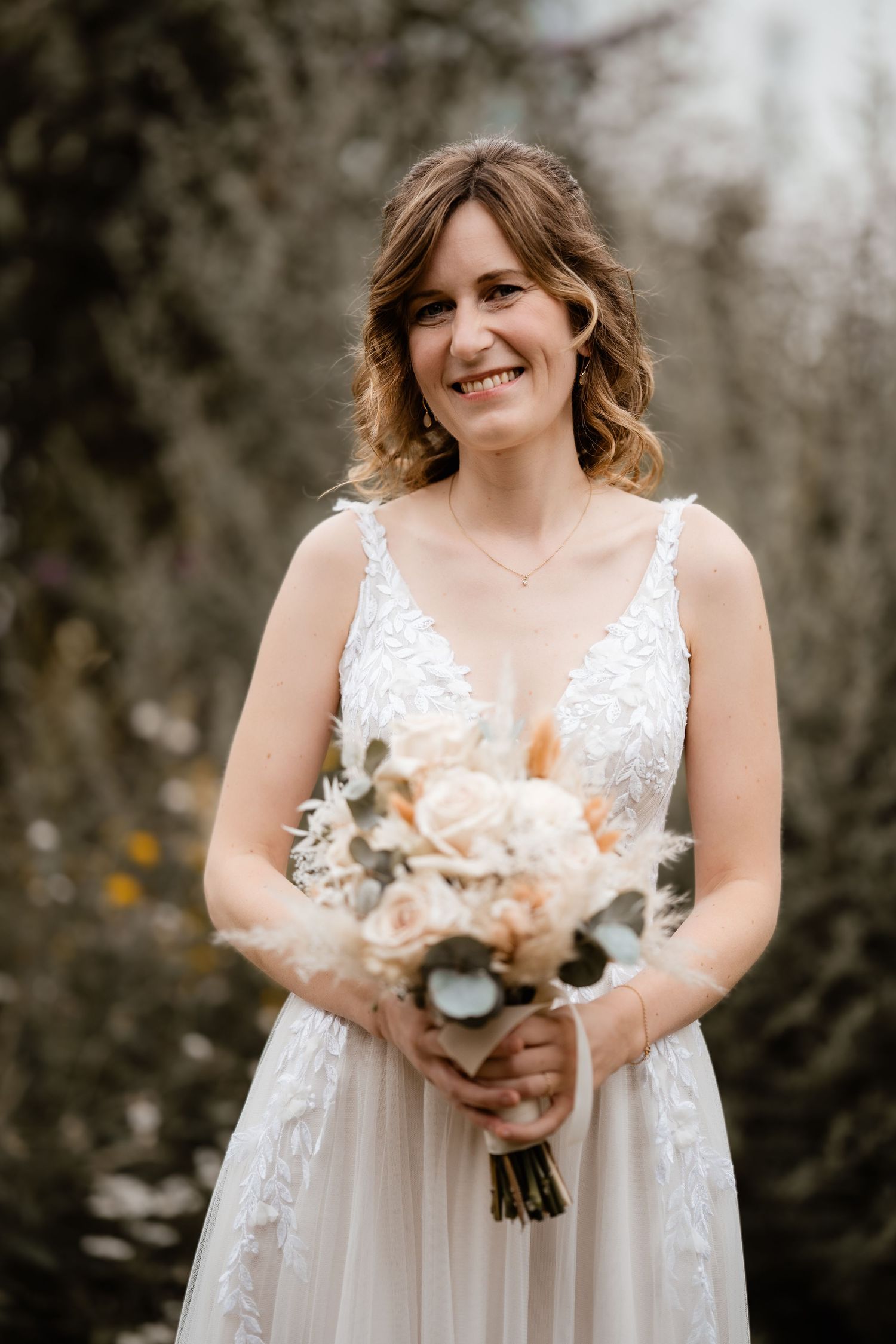 Smiling bride in a V-neck lace wedding dress holding a peach and white floral bouquet outdoors.