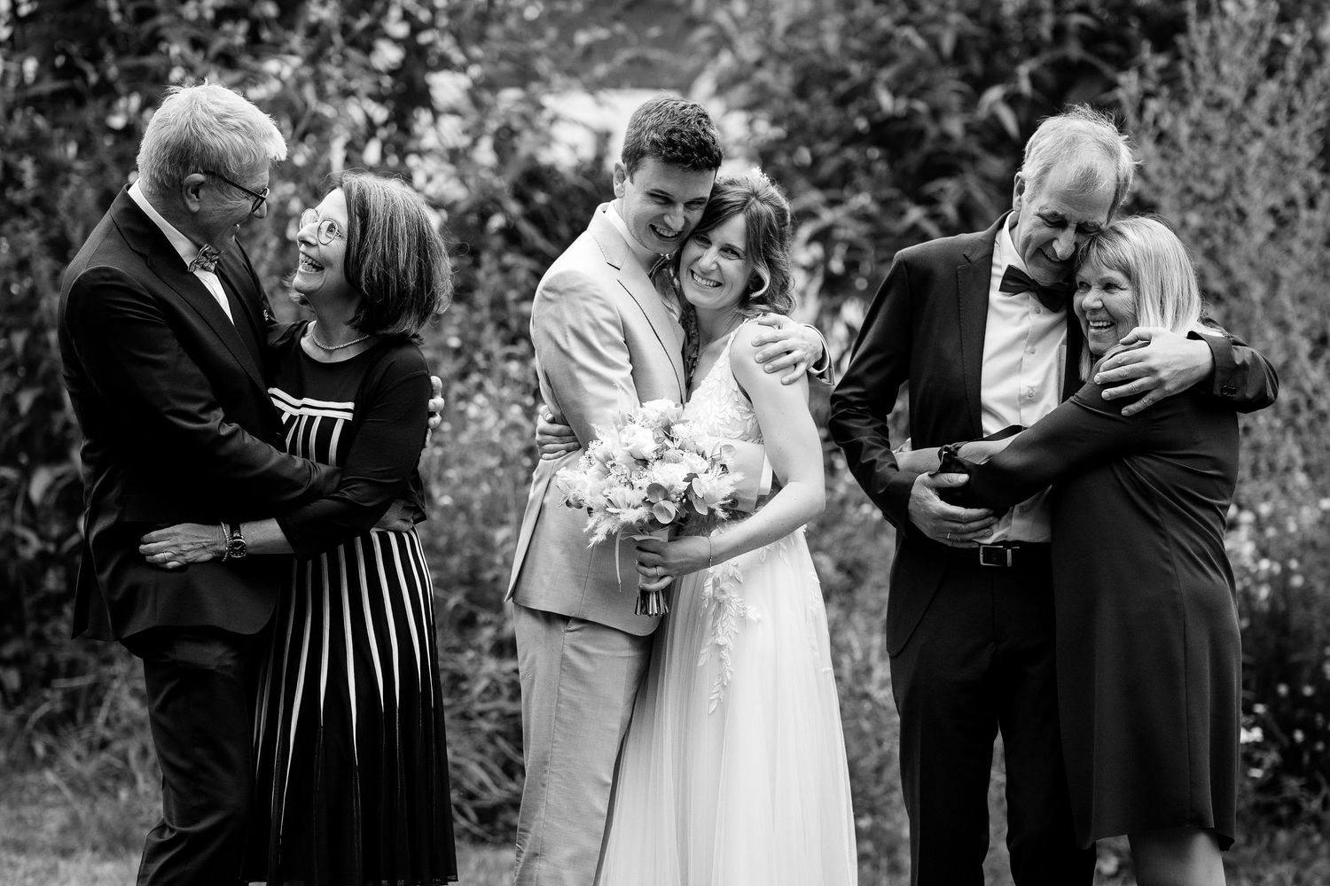 A newlywed couple embraces with both sets of parents in a joyful black and white family portrait.