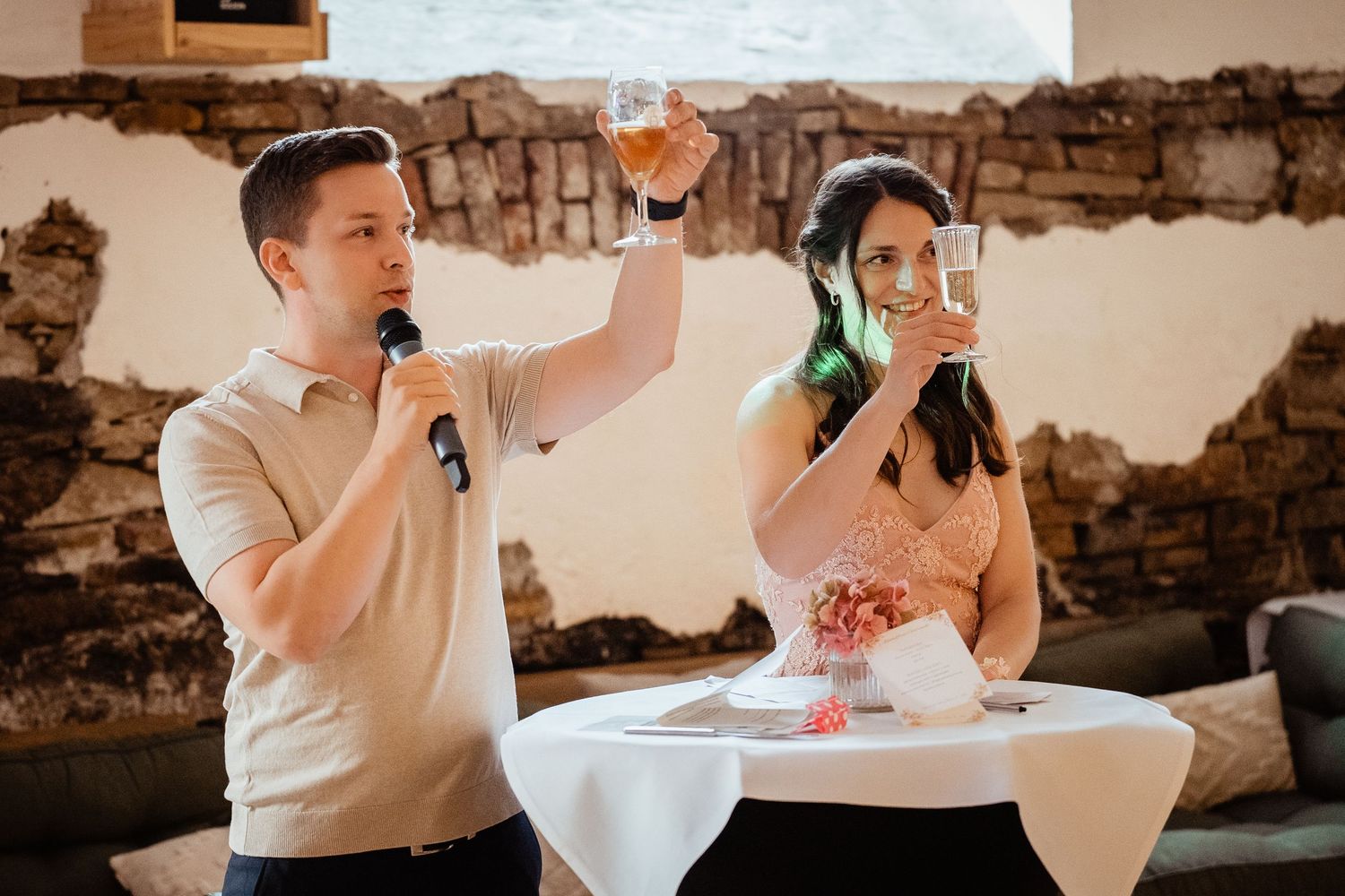 Man with microphone toasting alongside woman holding champagne glass at wedding reception.