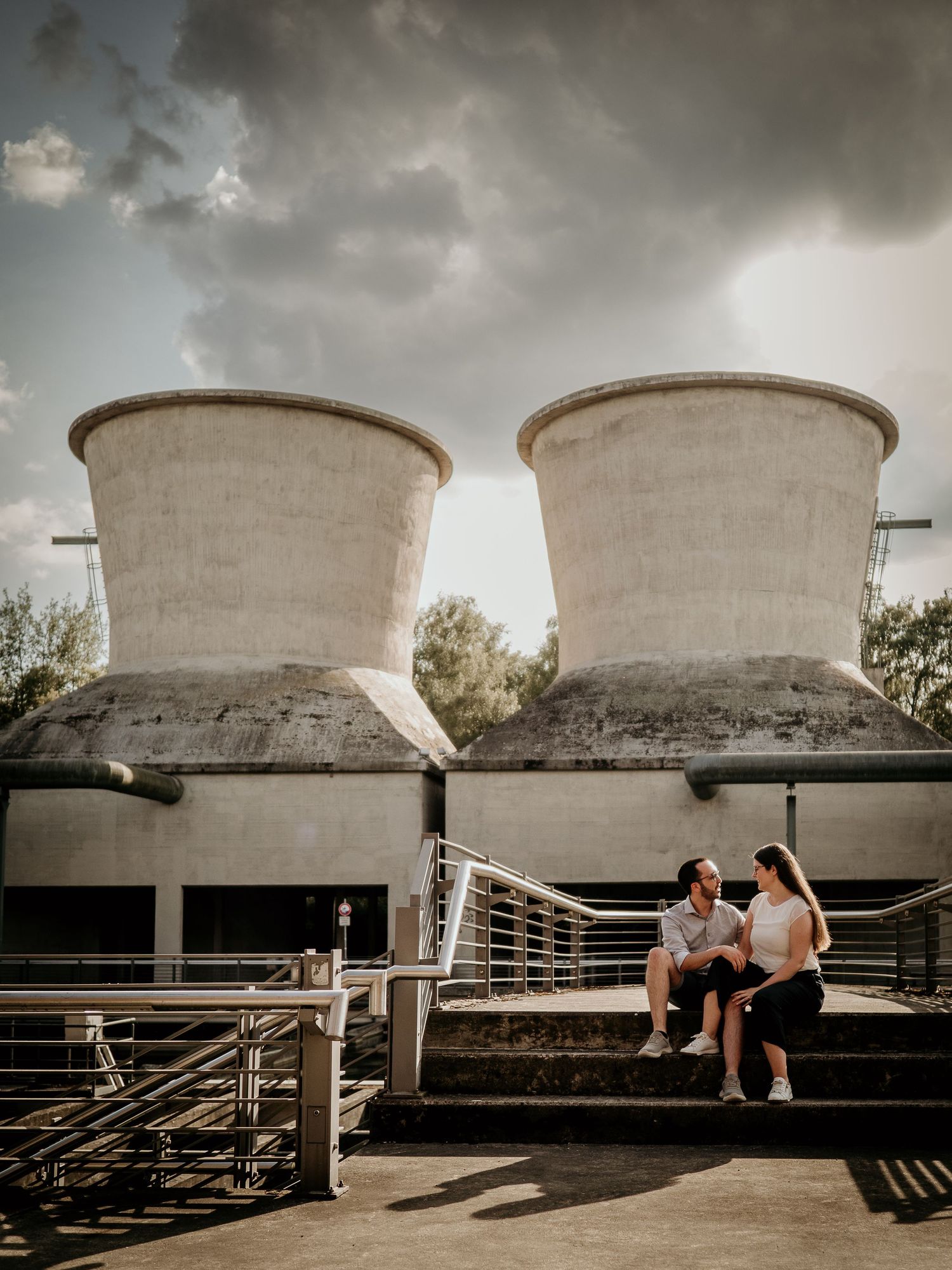 Couple sitting on steps in front of two large concrete cooling towers under dramatic cloudy sky.