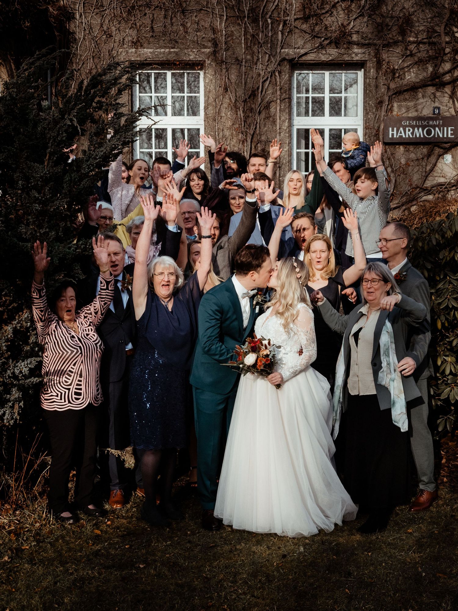 Wedding couple kisses surrounded by cheering guests outside Harmonie venue in autumn setting.