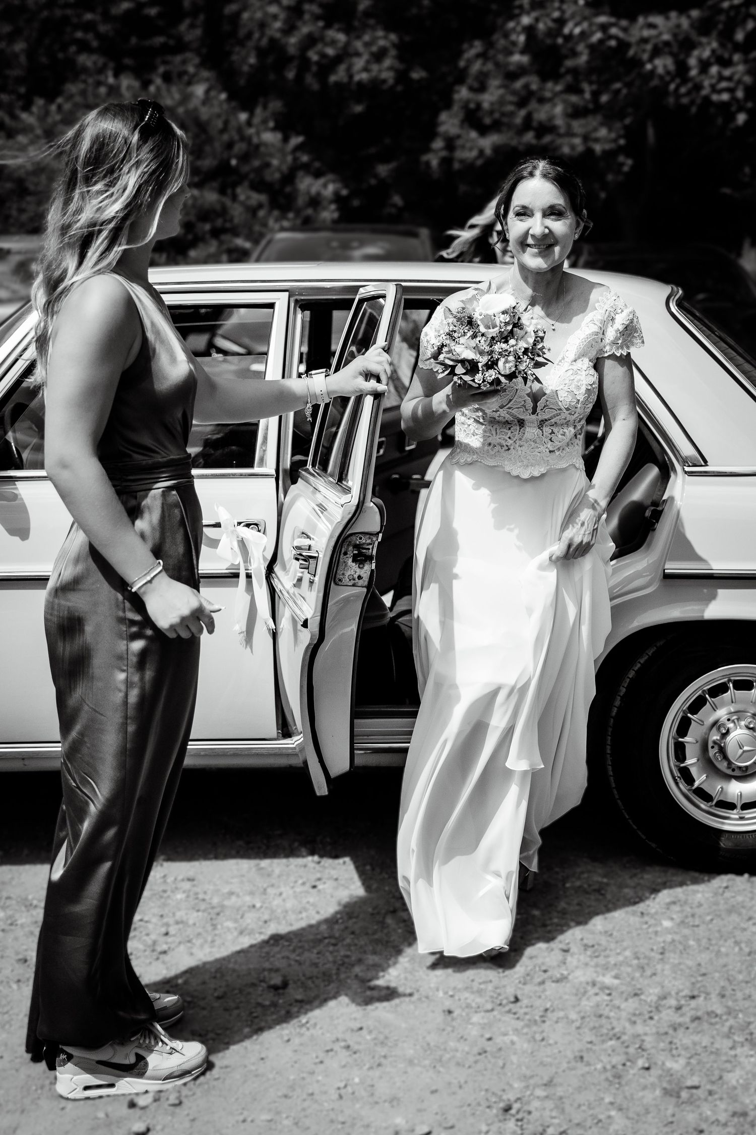 Bride in white lace gown holding bouquet steps out of vintage car assisted by bridesmaid in black outfit.