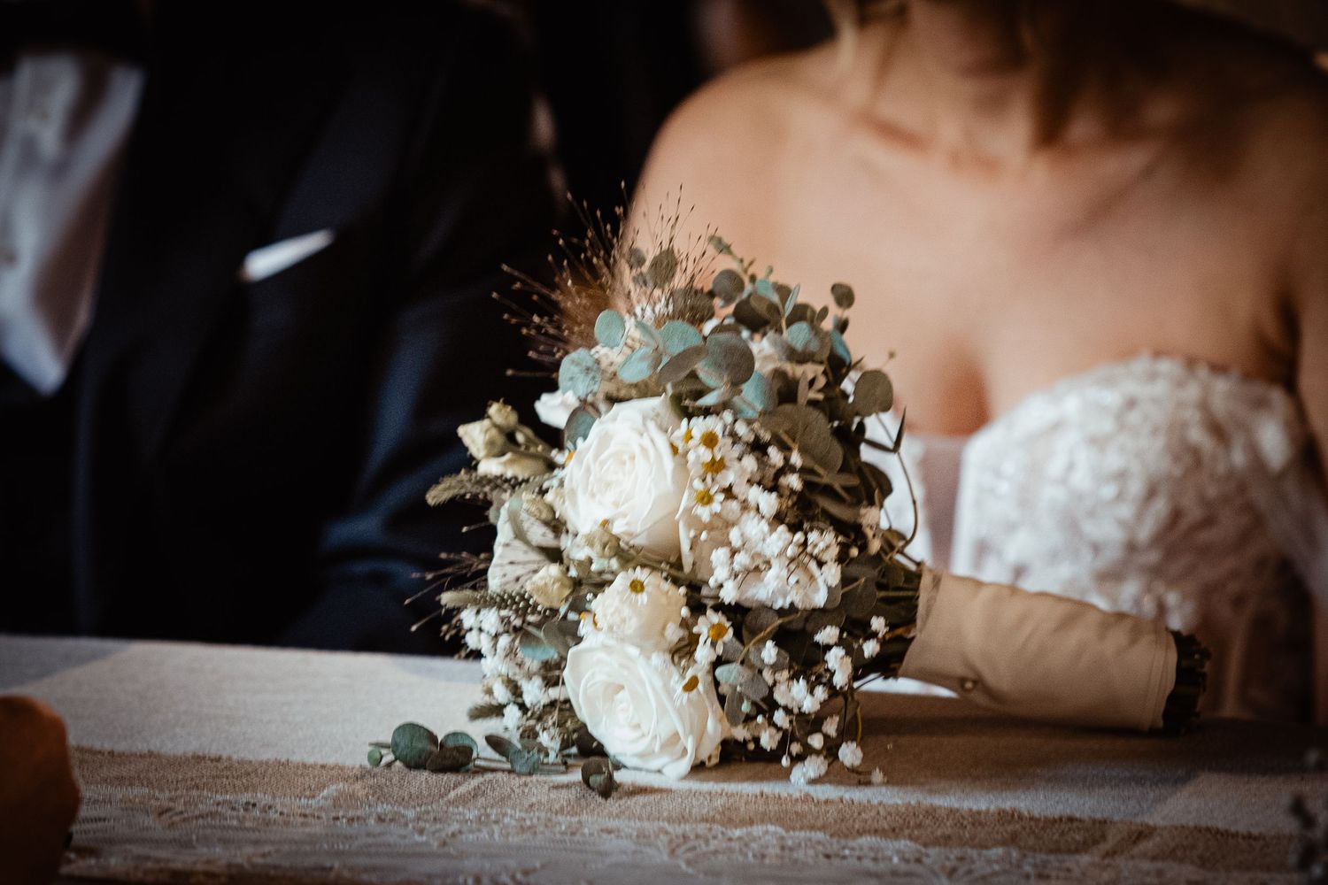 Bride holding a elegant wedding bouquet of white roses, eucalyptus, and dried flowers on a table.