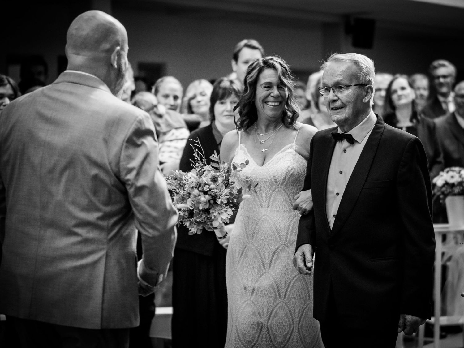 Black and white wedding ceremony photo of bride in lace gown walking down aisle with older gentleman.