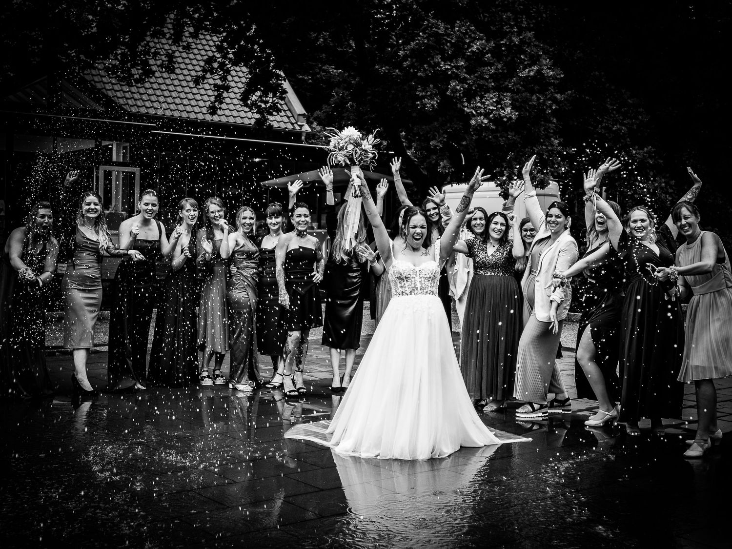 Black and white wedding photo of bride and groom celebrating with guests in rain, arms raised joyfully.