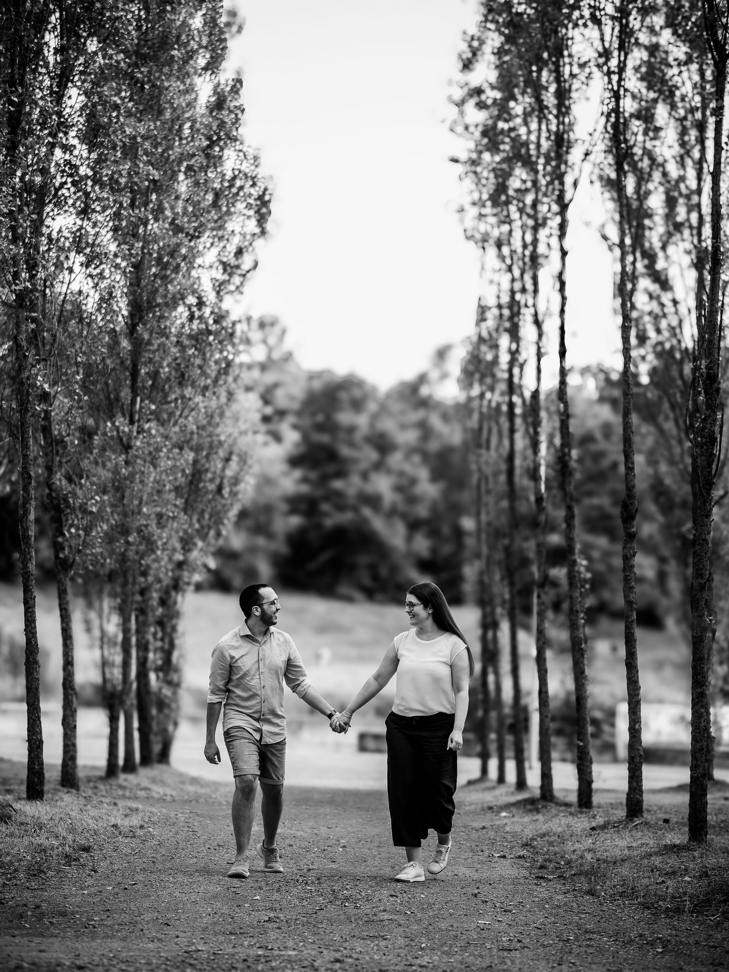 Couple holding hands and walking along a tree-lined path in a romantic black and white outdoor portrait.