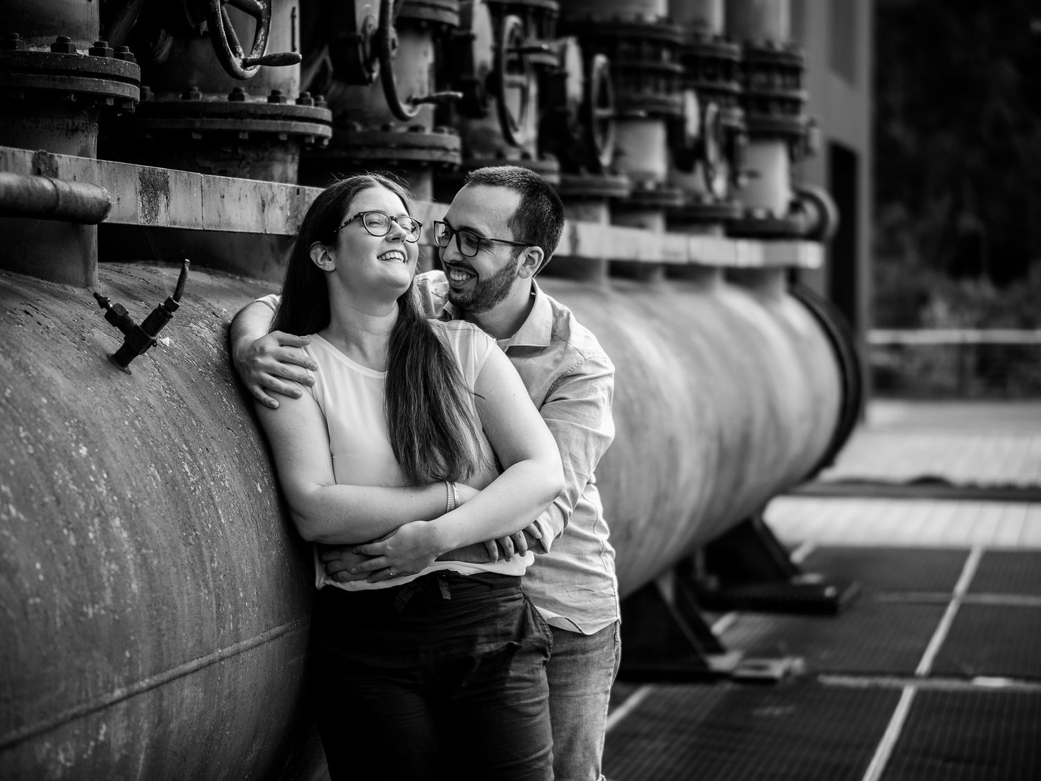 Black and white photo of a smiling couple embracing near large industrial pipes outdoors.