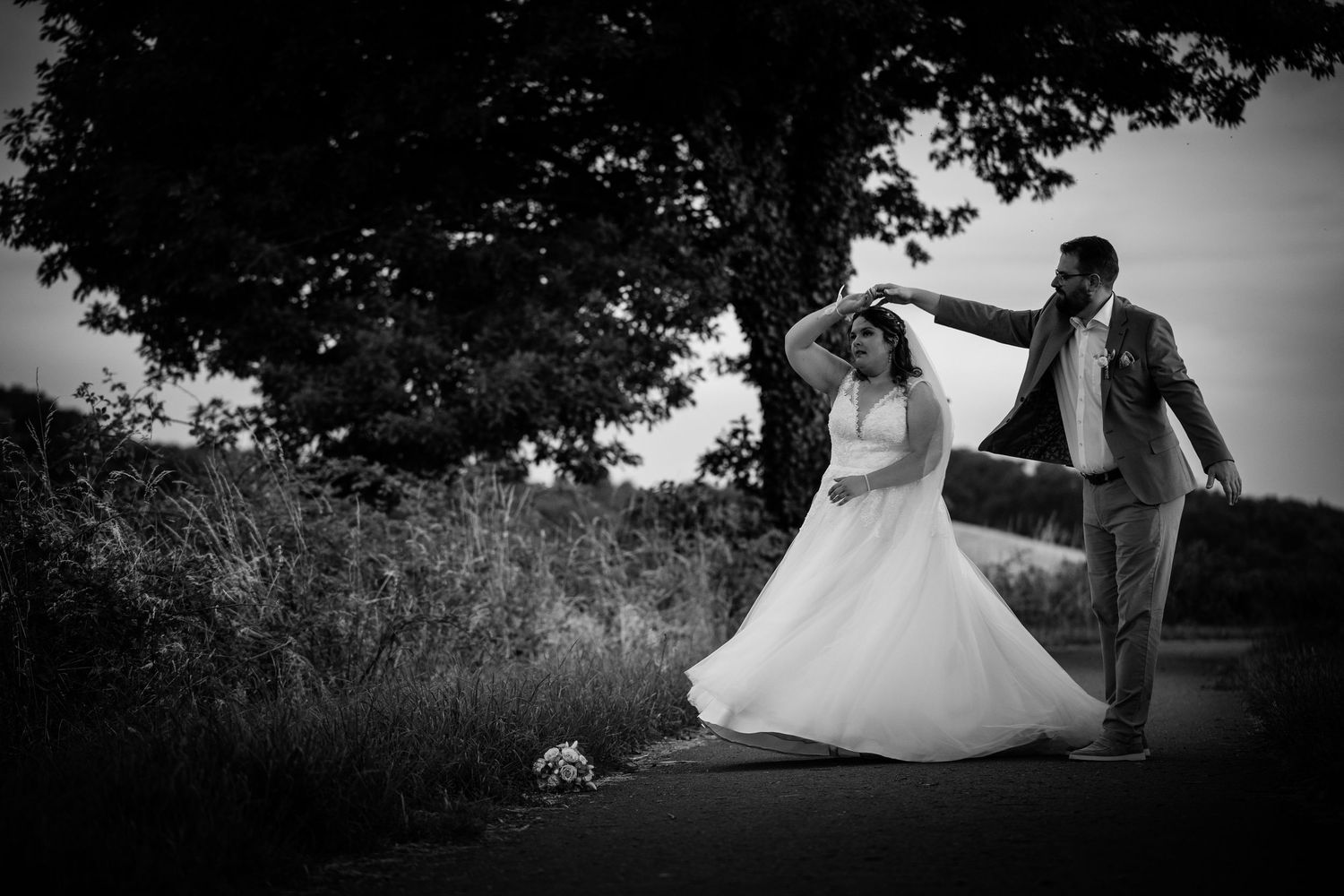 Black and white photo of groom twirling bride on country road beneath large tree at sunset.