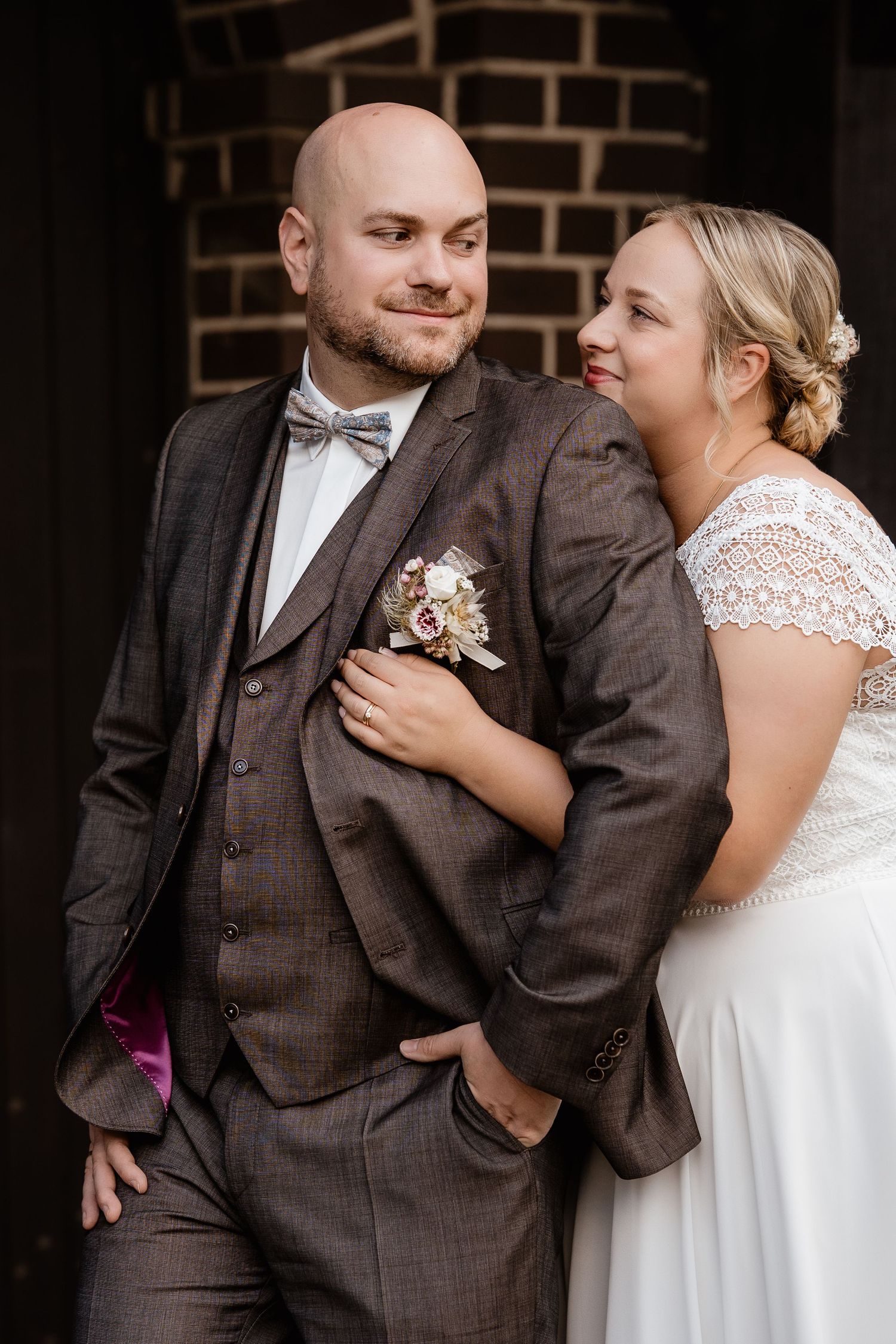 Bride in white lace dress leans on groom in brown three-piece suit with boutonniere, posing before brick wall.