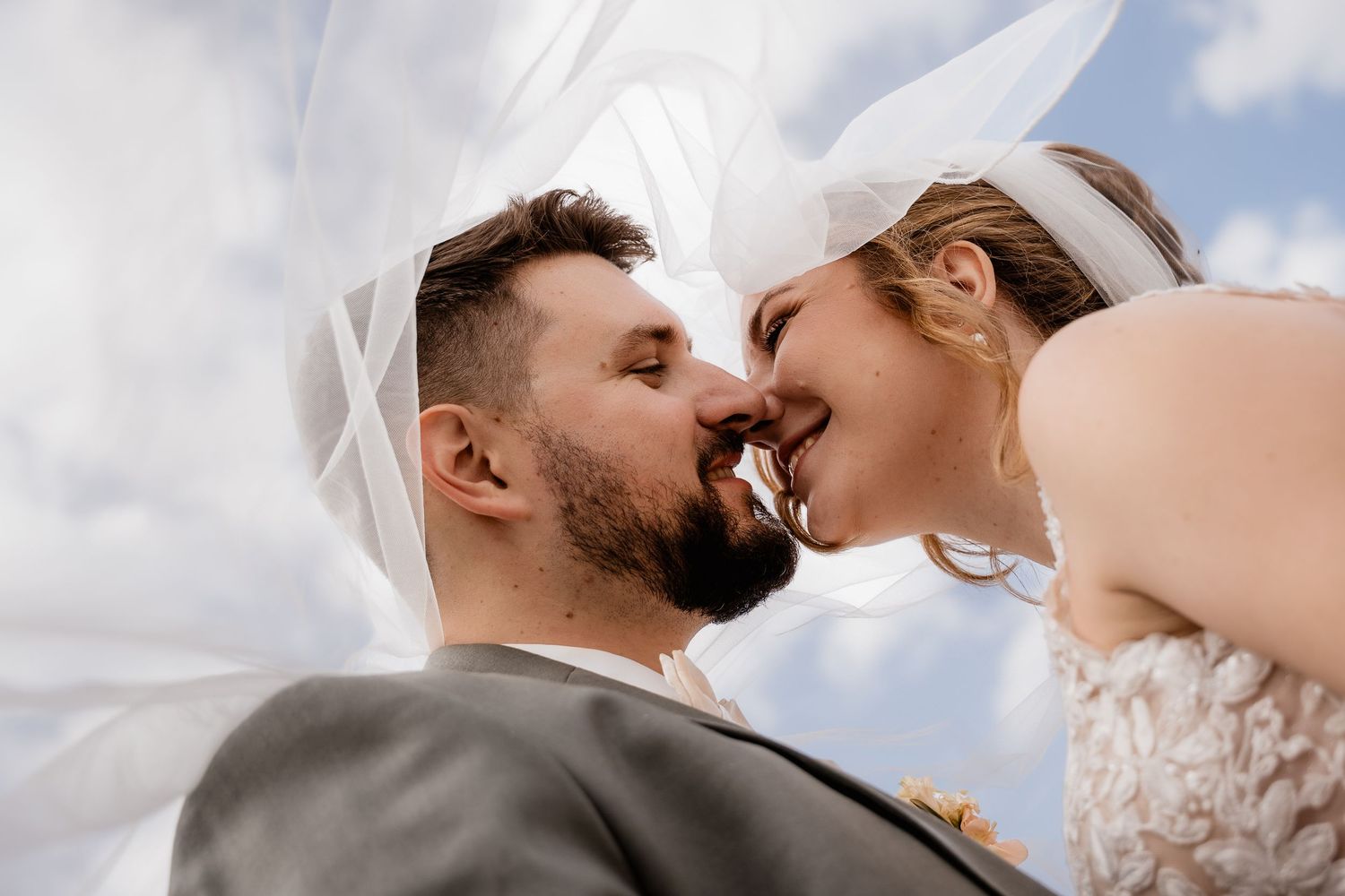 Bride and groom sharing a romantic kiss under flowing white veil against a blue sky on their wedding day.