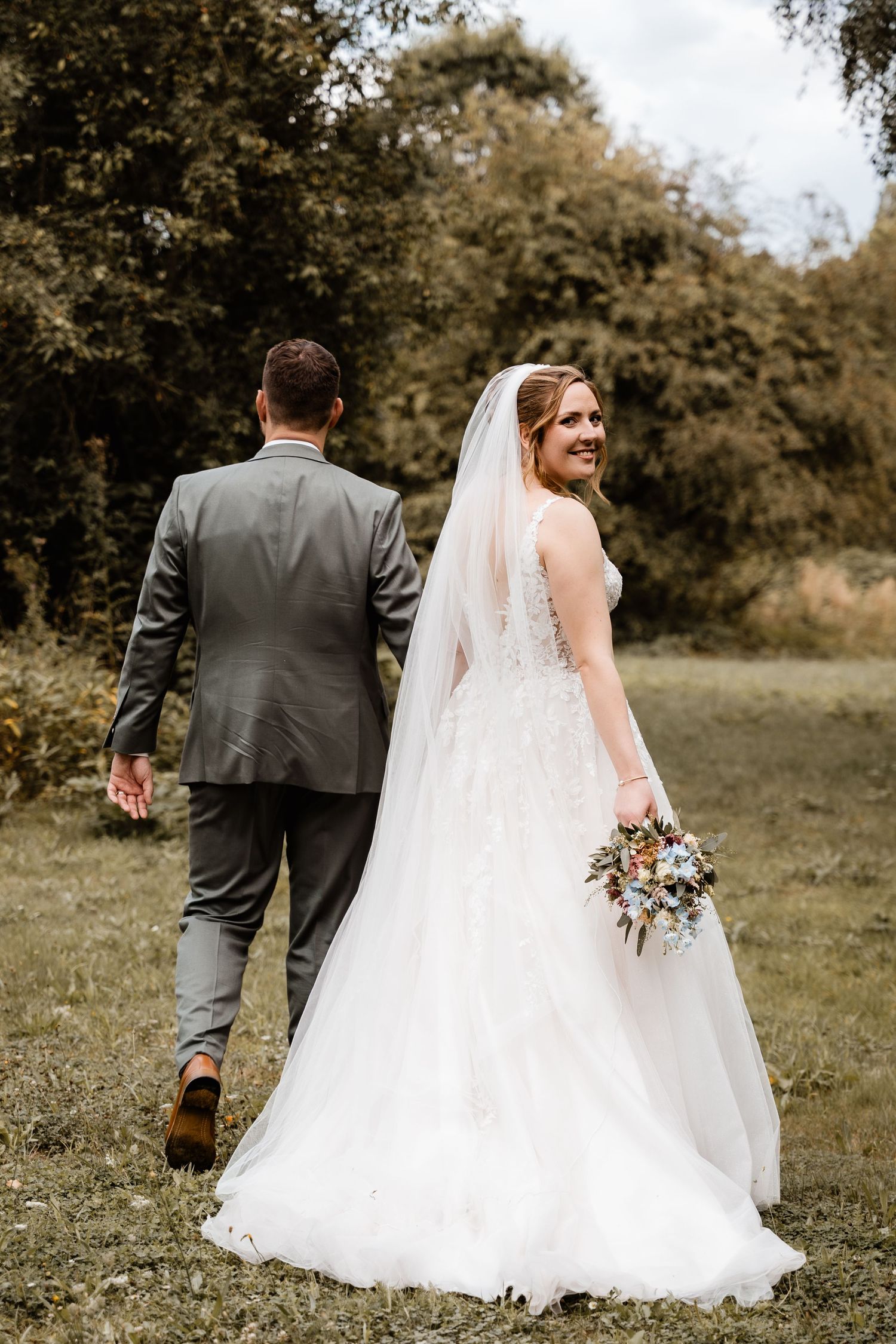 Bride in white lace gown with veil holds bouquet, glancing back while walking with groom in gray suit outdoors.