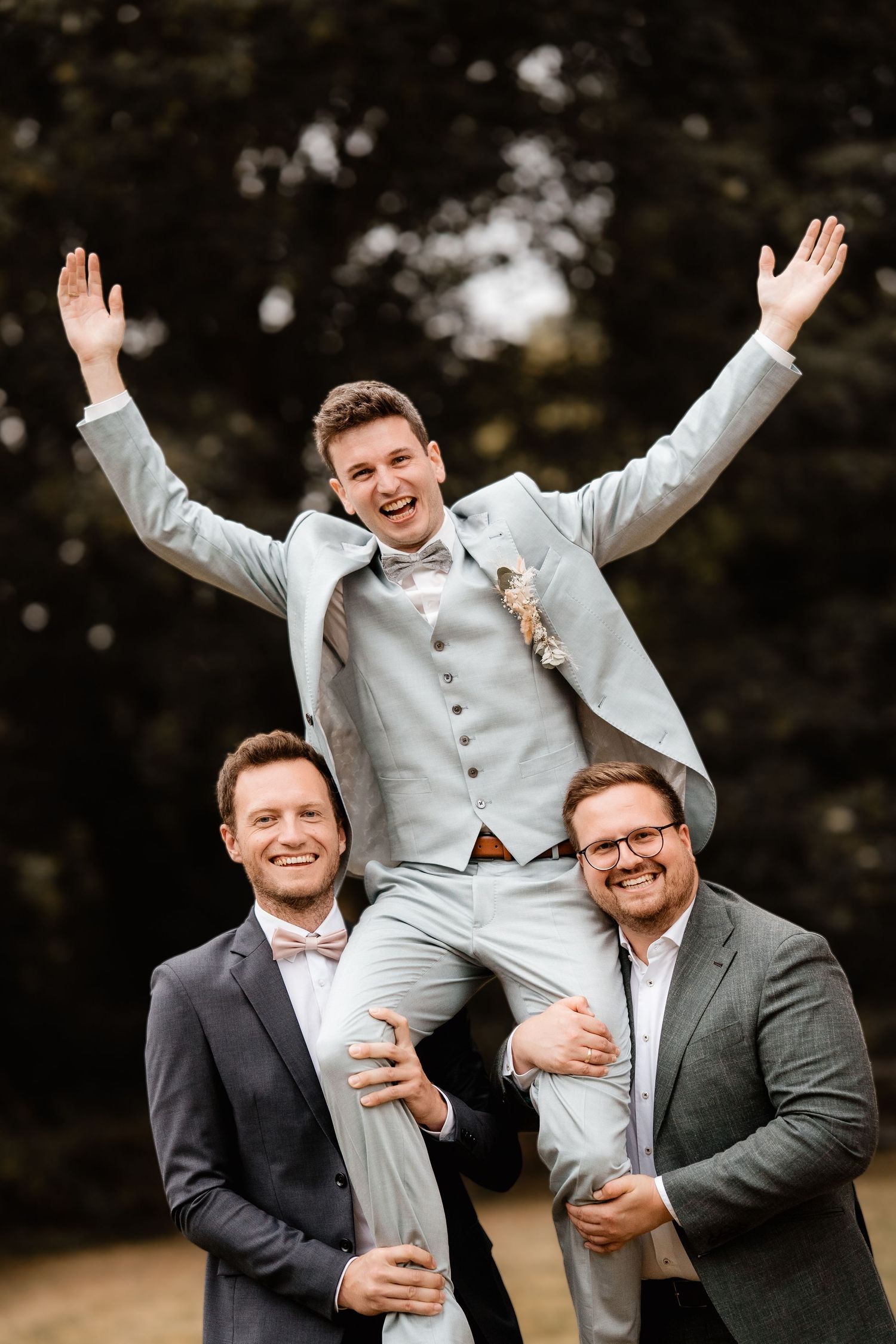 Excited groom with arms raised sits on shoulders of two smiling groomsmen outdoors.