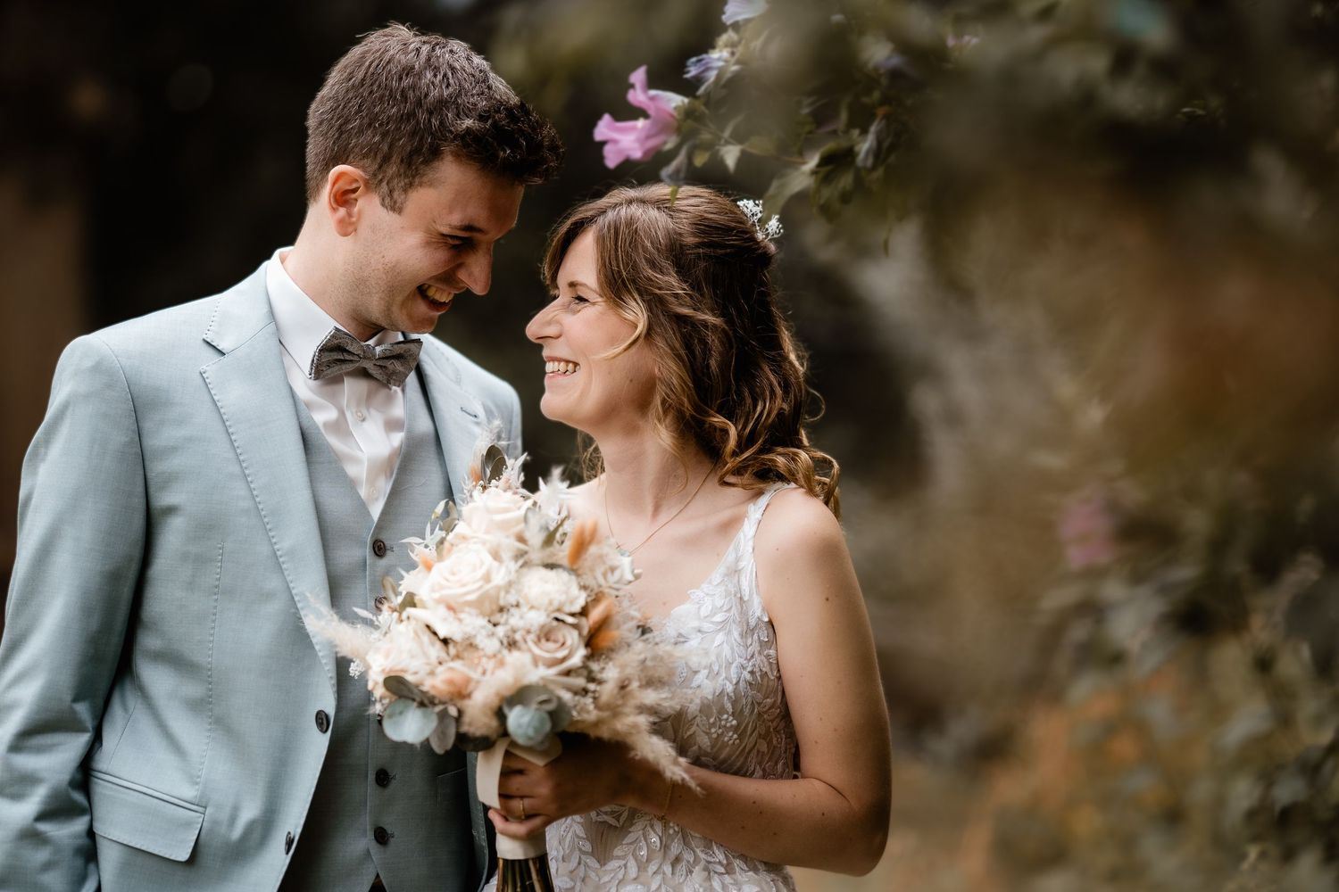 Bride and groom in light gray suit smile at each other holding a boho bouquet outdoors.