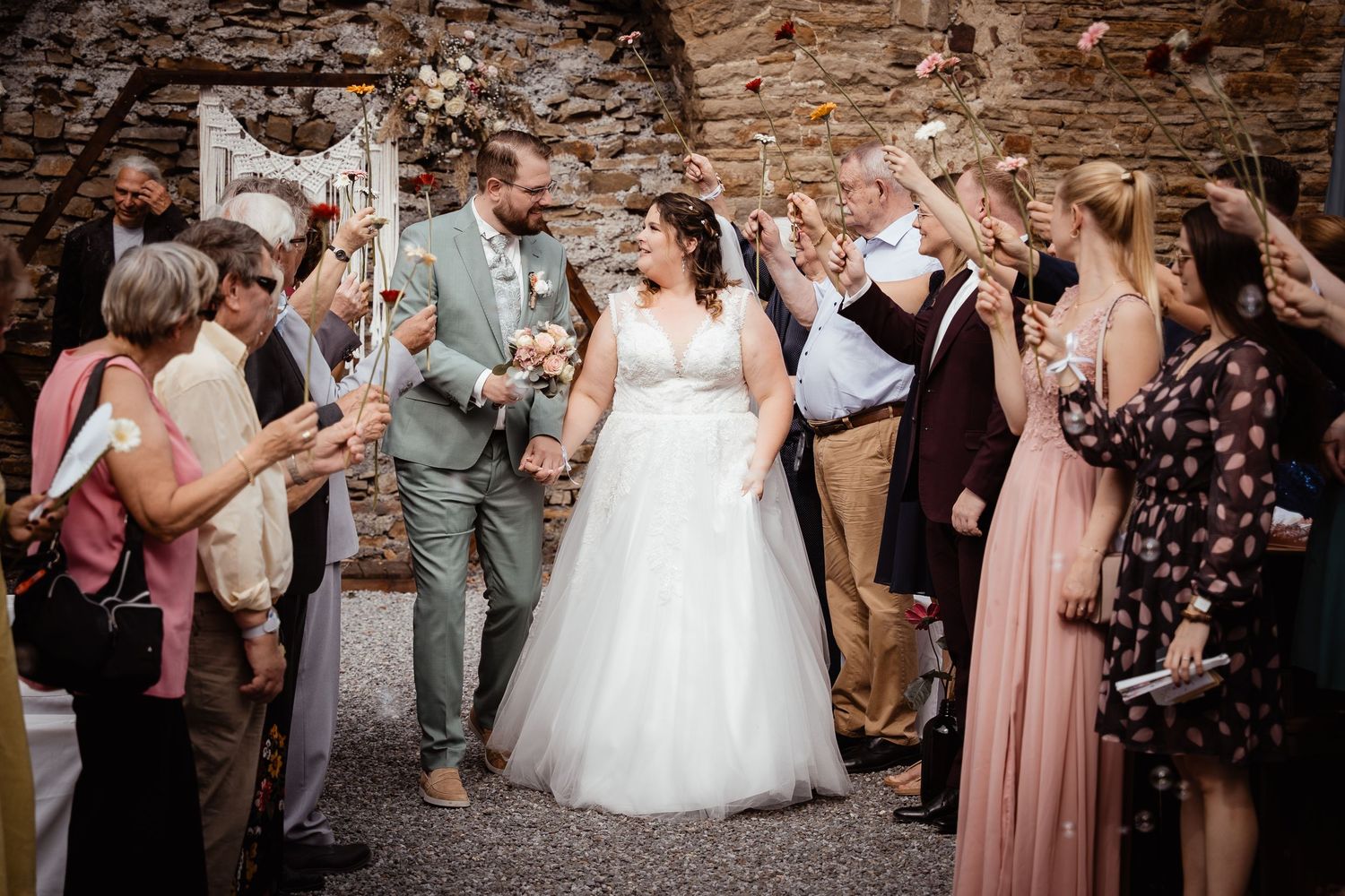 Newlyweds walk through a joyful confetti shower surrounded by cheering wedding guests in a stone venue.