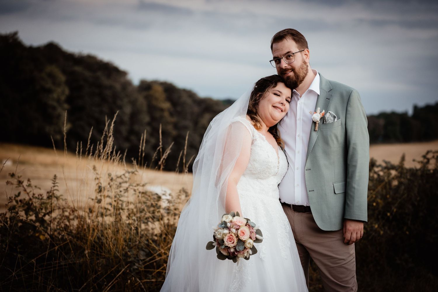 Bride and groom embracing in golden field with bouquet of roses and dramatic cloudy sky backdrop.