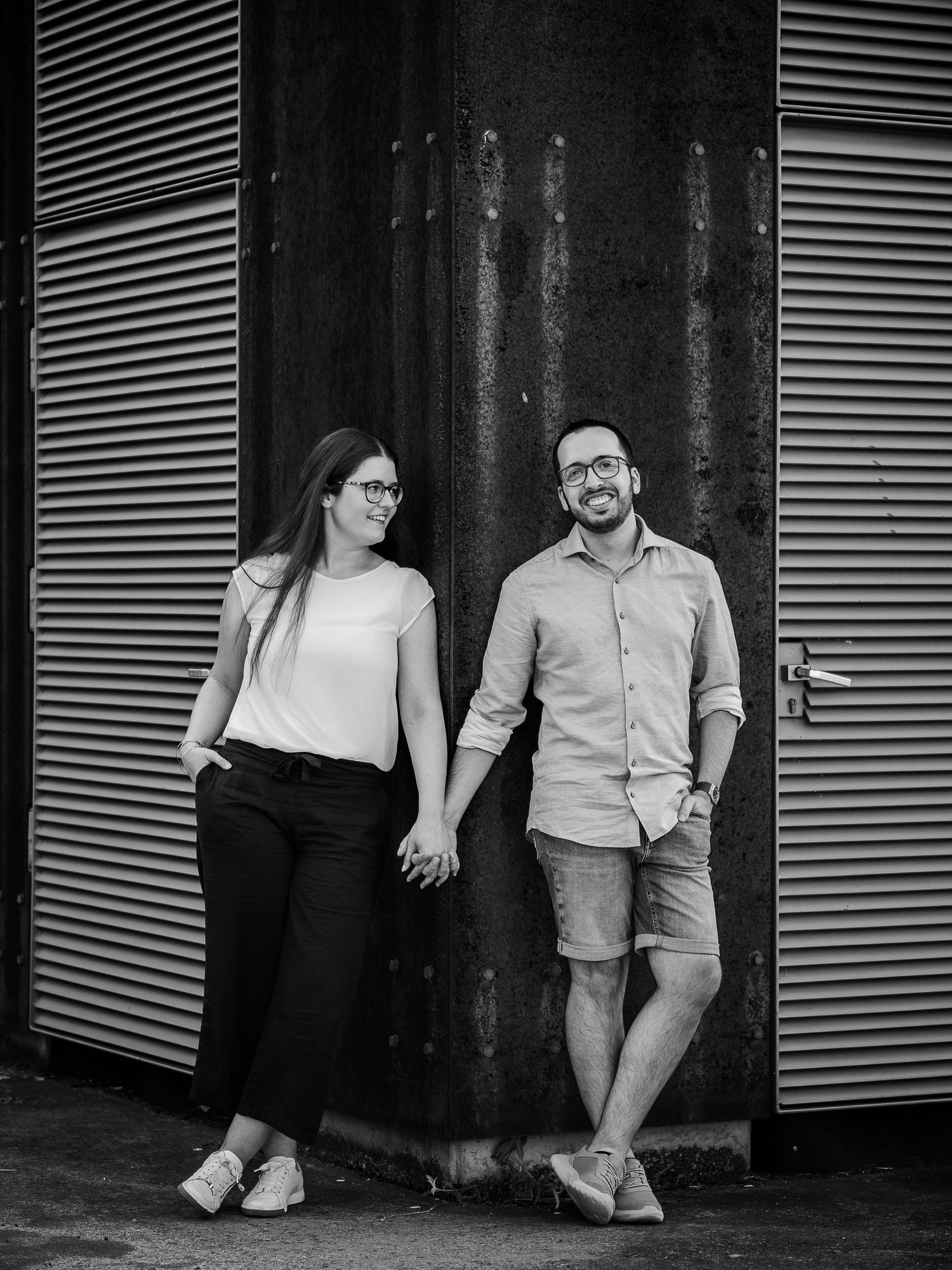 Black and white portrait of a smiling couple holding hands while leaning against corrugated metal doors.