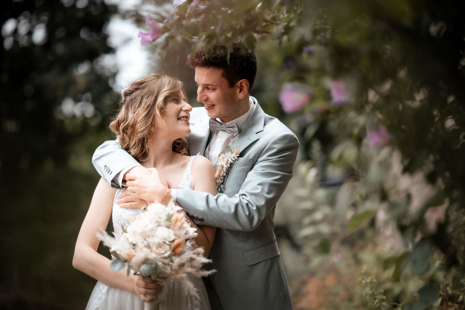 A smiling bride holding a bouquet embraces her groom in a gray suit outdoors among blooming flowers.