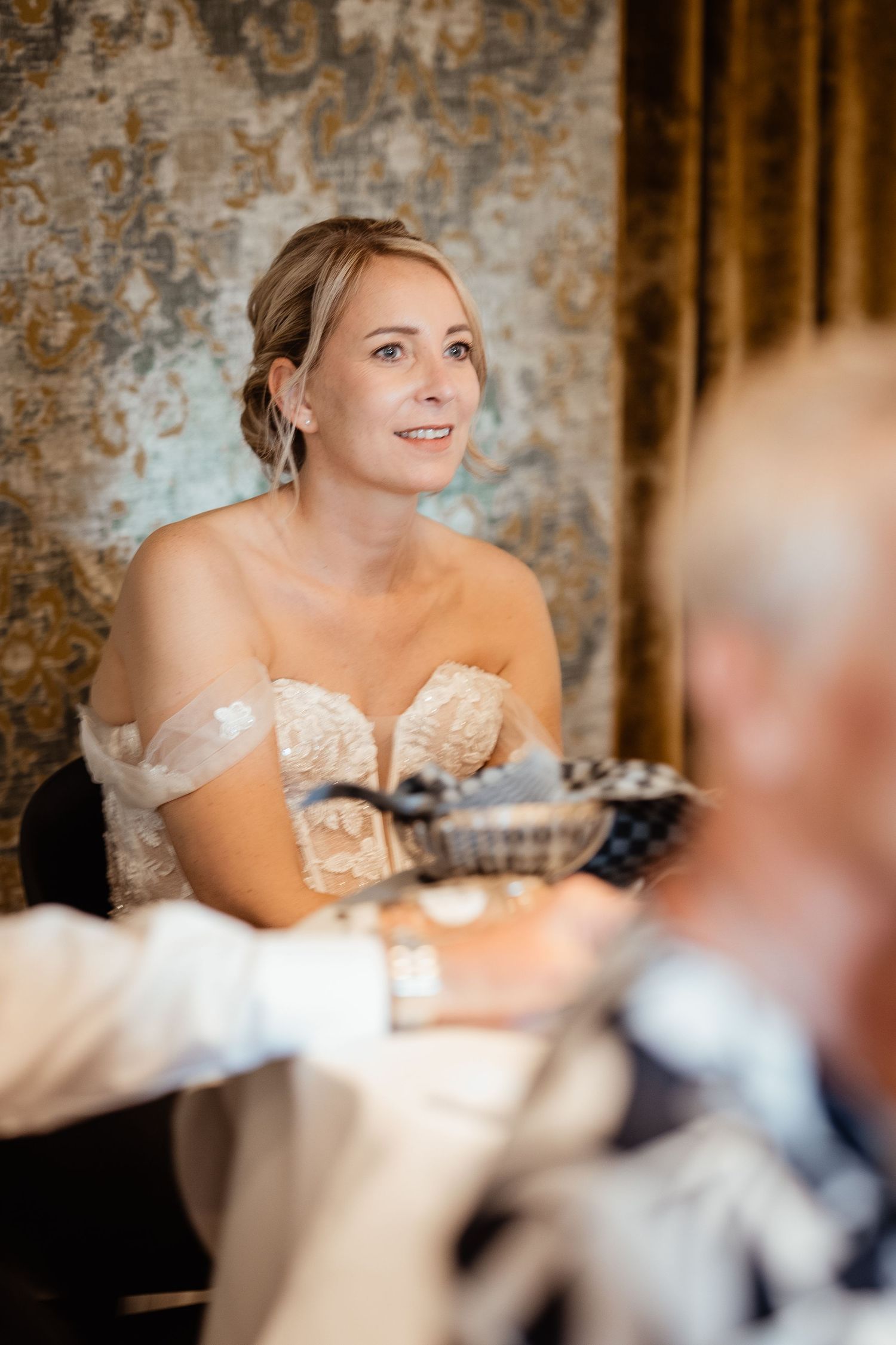 Elegant bride in off-shoulder lace gown listening attentively during wedding reception speeches.